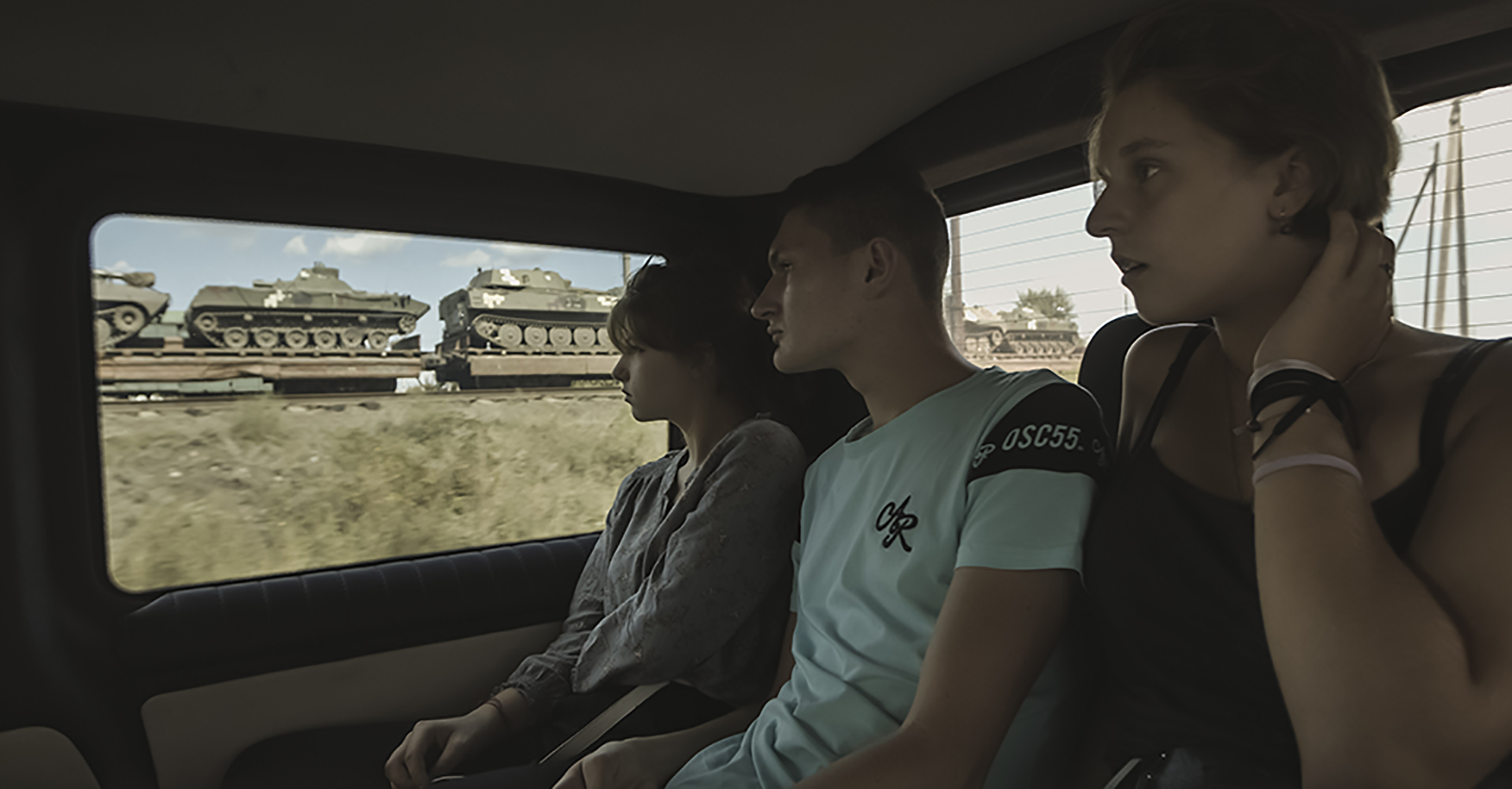 Tři mladí lidé sedí na zadním sedadle auta a sledují, jak se po kolejích vedle nich převáží vojenské tanky.