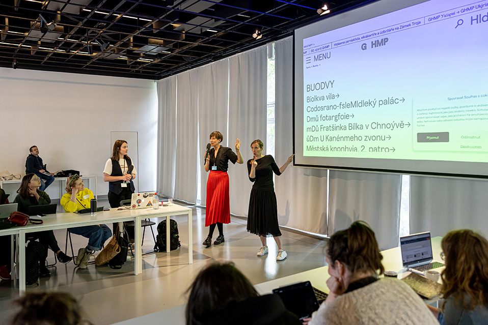 Seminář: Jak na přístupnou kulturu v CAMPu Ve velké moderní místnosti sedí v kruhu lidé u stolu a sledují prezentaci na projekčním plátně. U plána stojí osoba a překládá prezentaci do českého znakového jazyka.