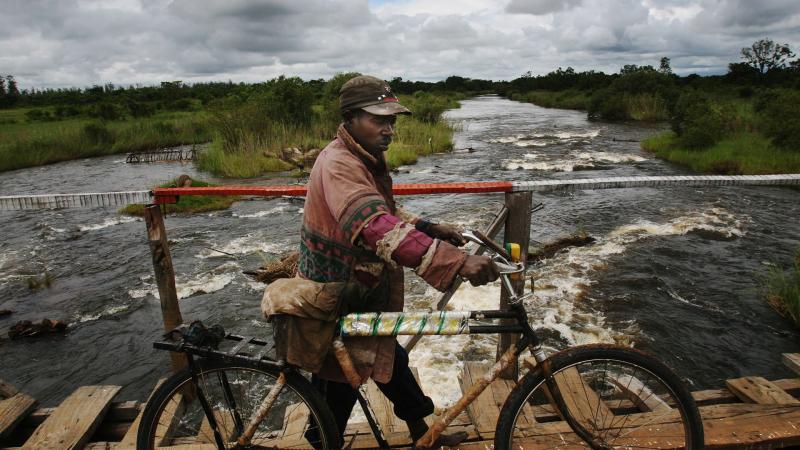 Muž vede různě opravované kolo přes chatraný dřevěný most přes řeku
