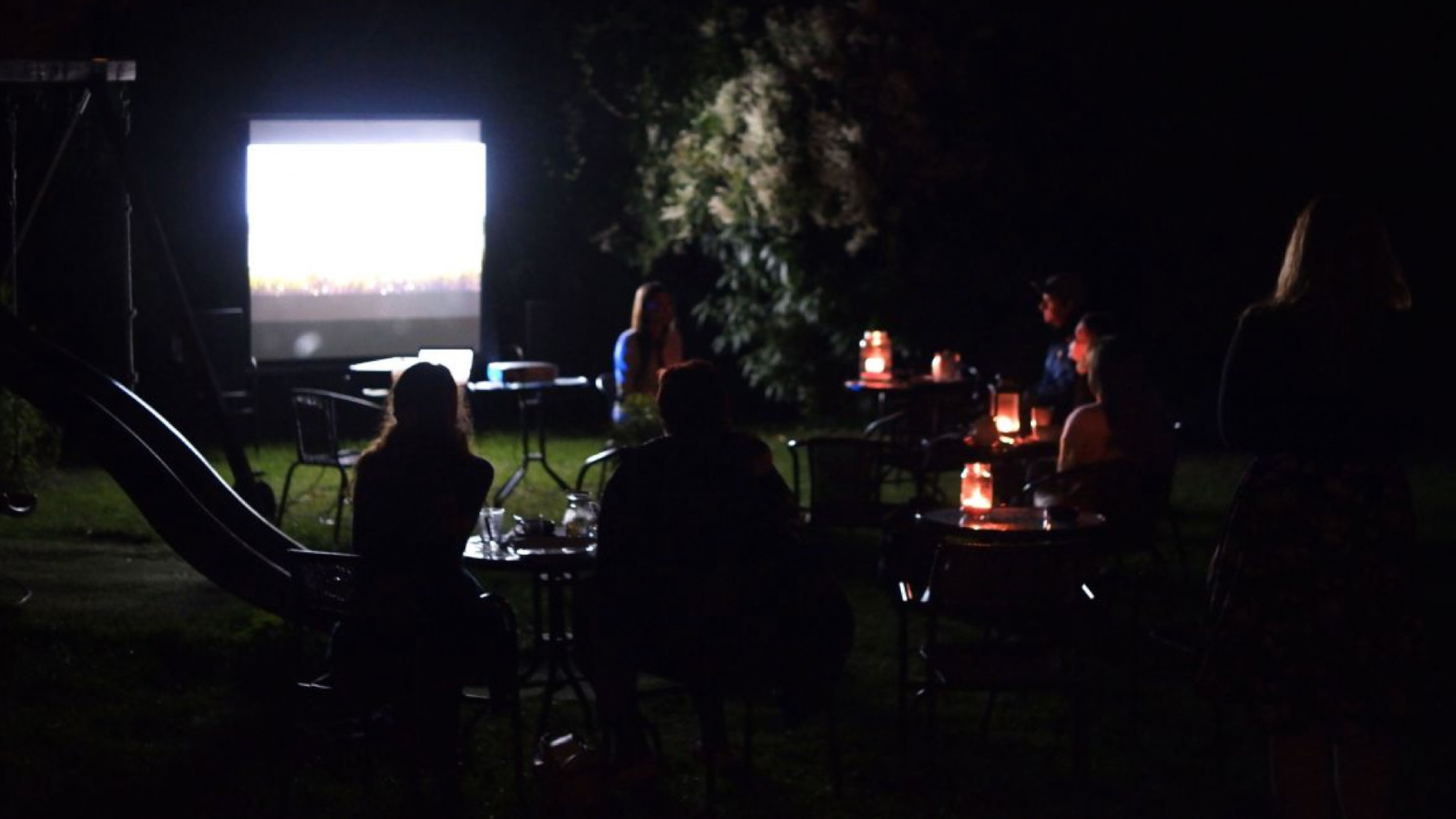 Promítání filmu večer na zahradě. Diváci sedí u stolečků se svíčkama.