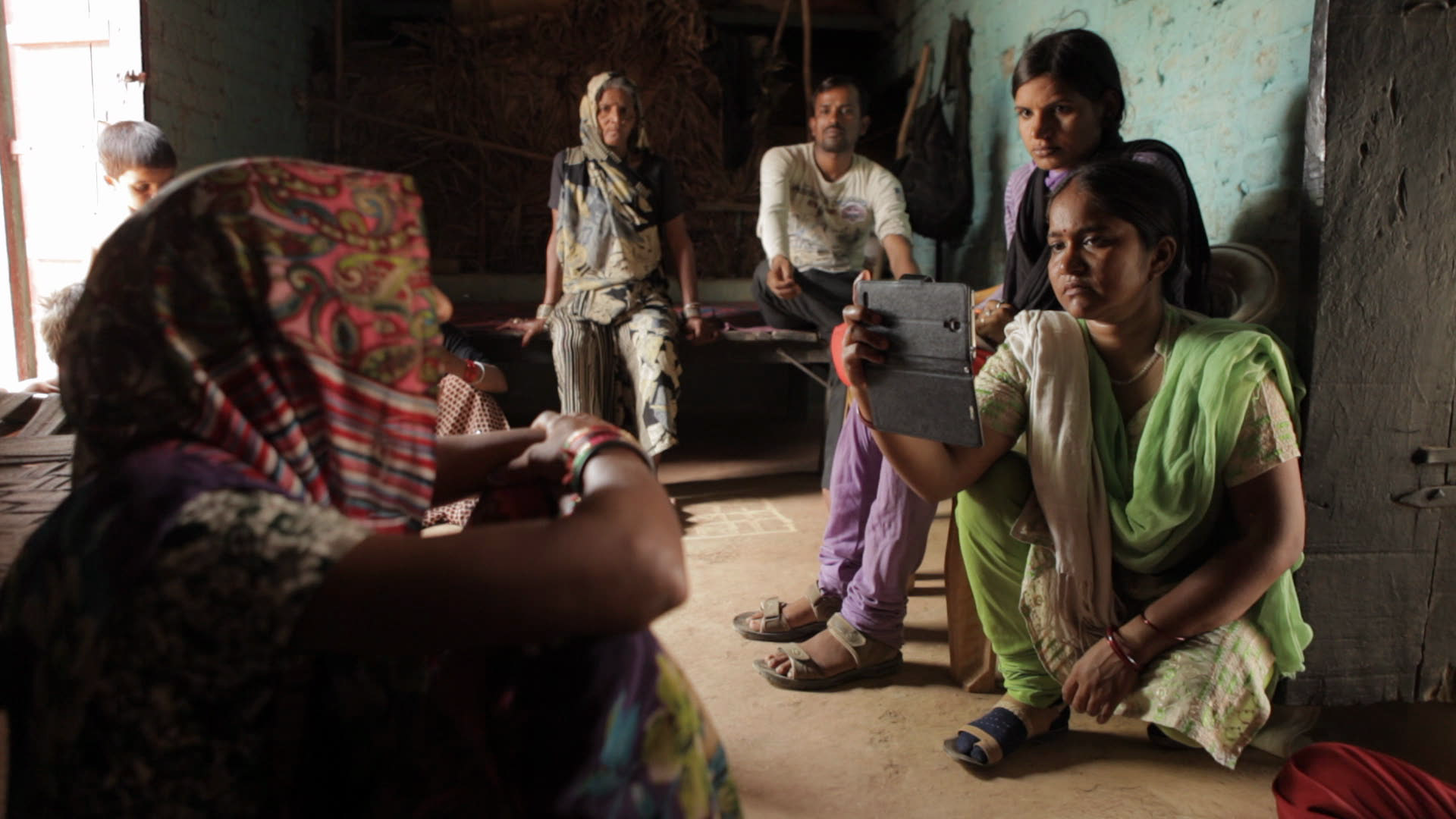Skupinka žen sedí na hliněné podlaze v přízemním domku. Jedna z nich nahrává video do mobilu.