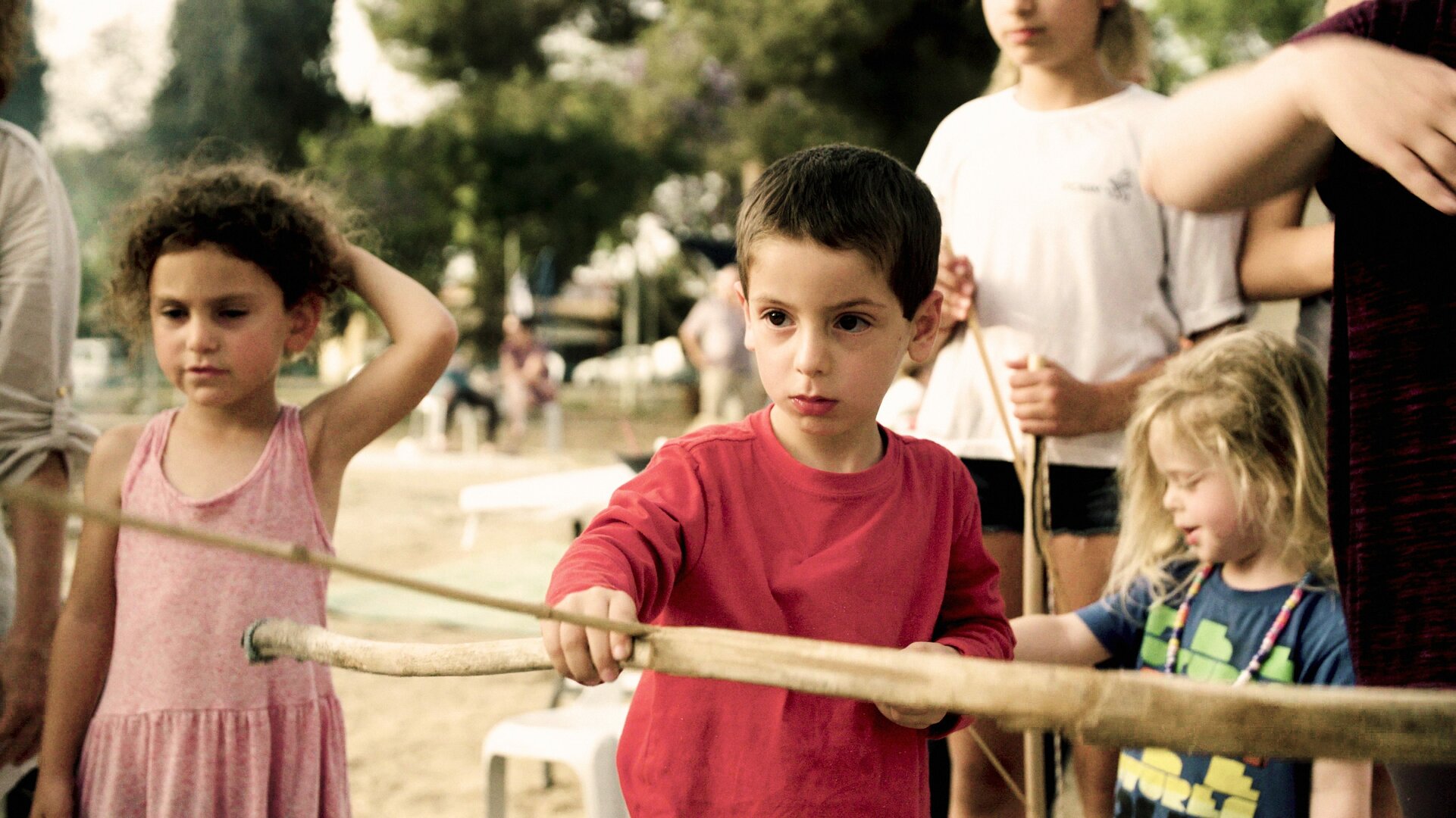 Chlapec na dětském hřišti se skupince děti drží v rukou dřevěný luk a šíp a míří před sebe.