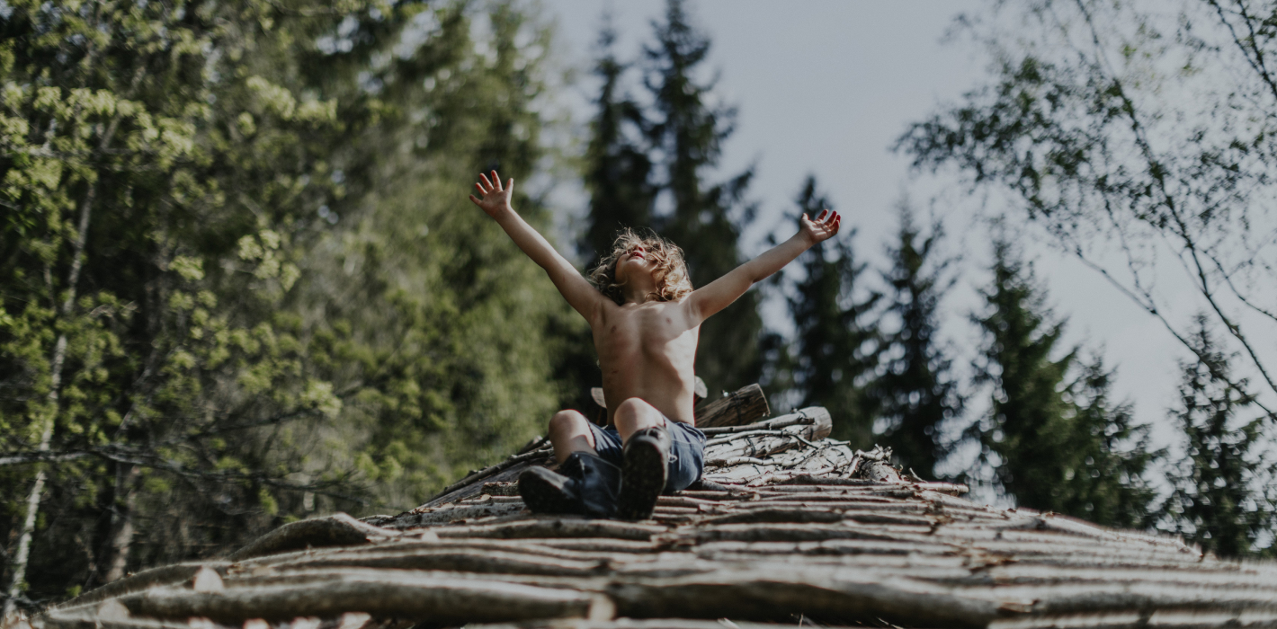 Chlapec s radostě zdviženýma rukama sedí na hromadě dříví v lese. / Child with arms raised sitting atop a wooden structure in the forest.