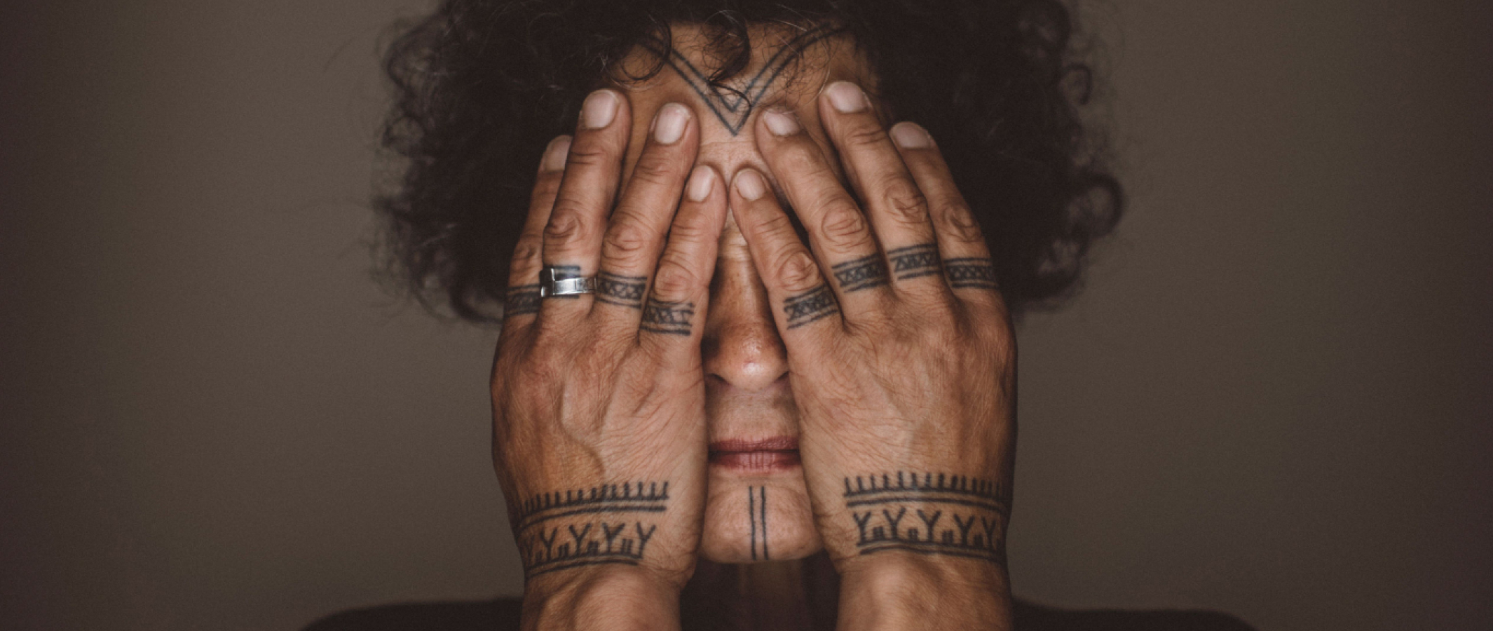 Osoba s tetováním na prstech si zakrývá obličej rukama. / Person with tattooed hands covering eyes, showing rings and detailed ink.