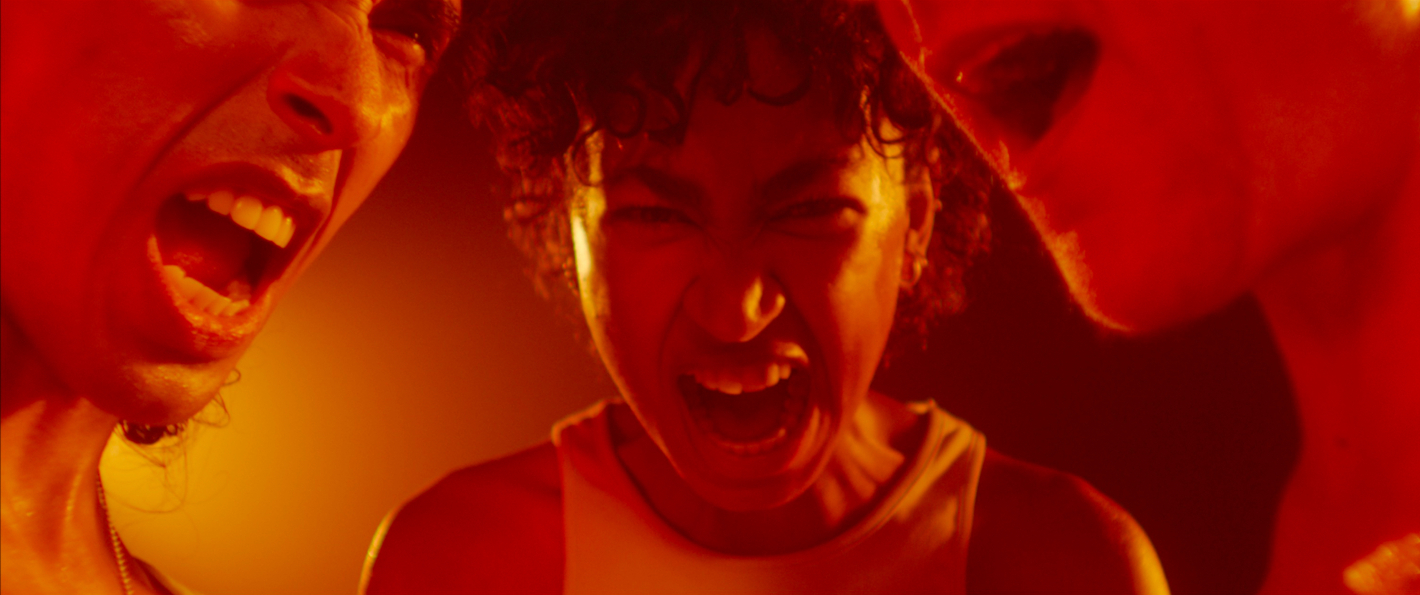 Tři rozlícené osoby křičí, červené osvětlení zdůrazňuje napětí. / Three people yelling in a red-tinted light.