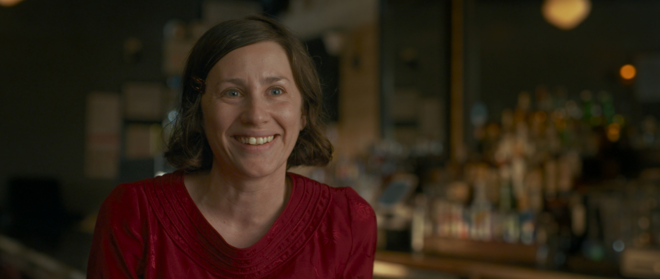 Žena se širokým úsměvem sedí v baru a mluví s člověkem za kamerou. / Smiling woman in red top with blurred bar background.