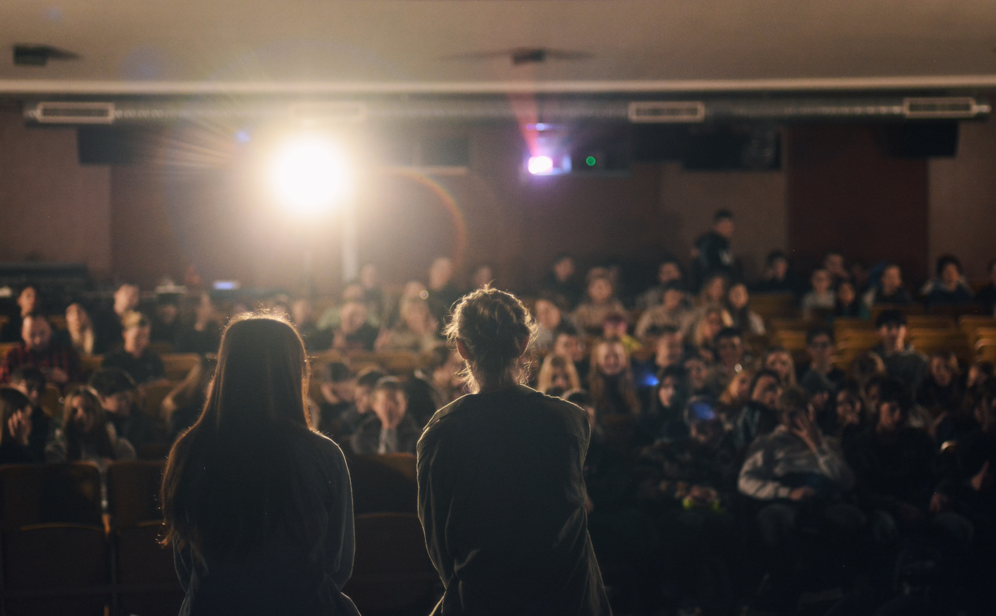 Dvě ženy sedí v popředí zády a debatují s diváky v plném kinosále.