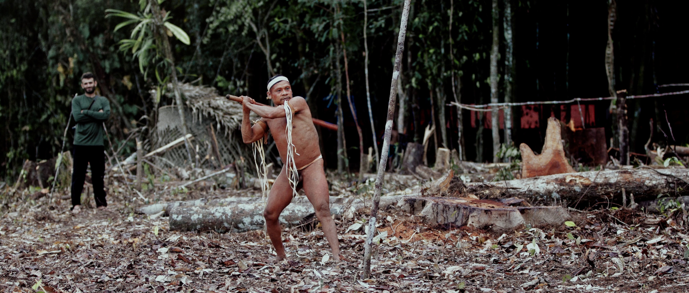 Muž v tradičním oděvu se rozmachuje s tyčí, druhý ho pozoruje ve vykáceném lese.