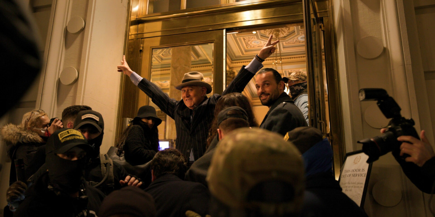 Muž v obleku zvedá ruce v pozdravu při východu z budovy, kolem je dav lidí a novinářů s kamerami.