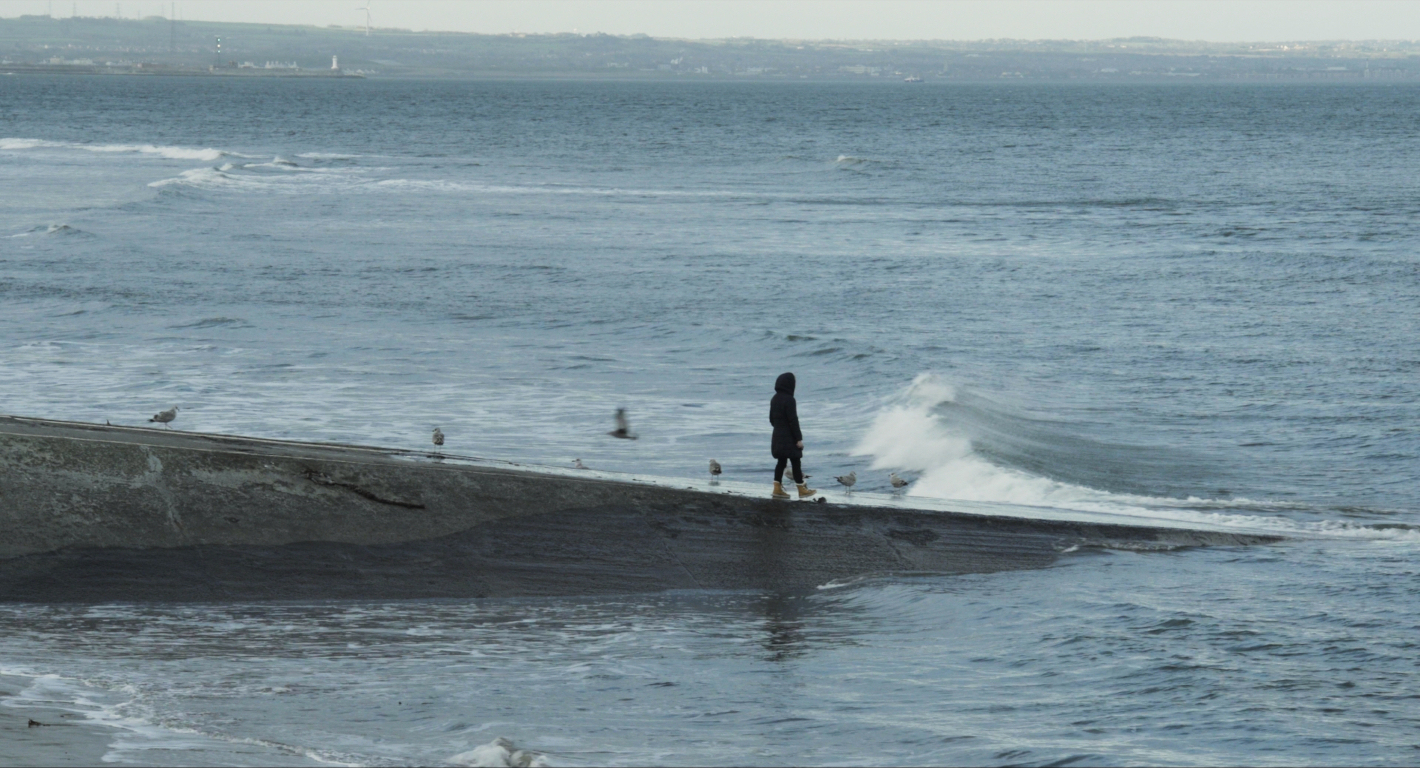 Uprostřed obrázku je žena stojící na molu, všude kolem moře.