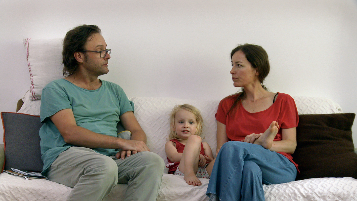 Muž a žena sedí na gauči a dívají se na sebe, mezi nimi sedí jejich malé dítě.