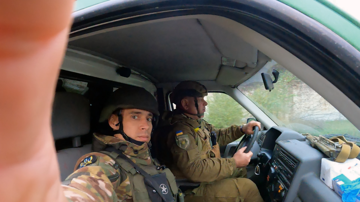 Dva ukrajinští vojáci ve vojenském oblečení s helmami jedou autem. Jeden z nich se dívá přímo do foťáku.