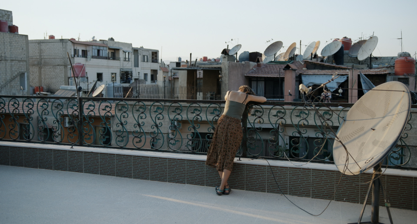Žena se opírá o zábradlí a je obrácená zády. Kolem jsou střechy domů poseté satelity. 