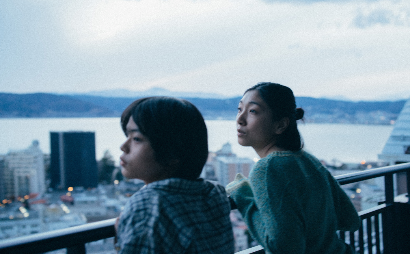 Dvě mladé dívky - asiatky - stojí na balkóně a dívají se do dálky.