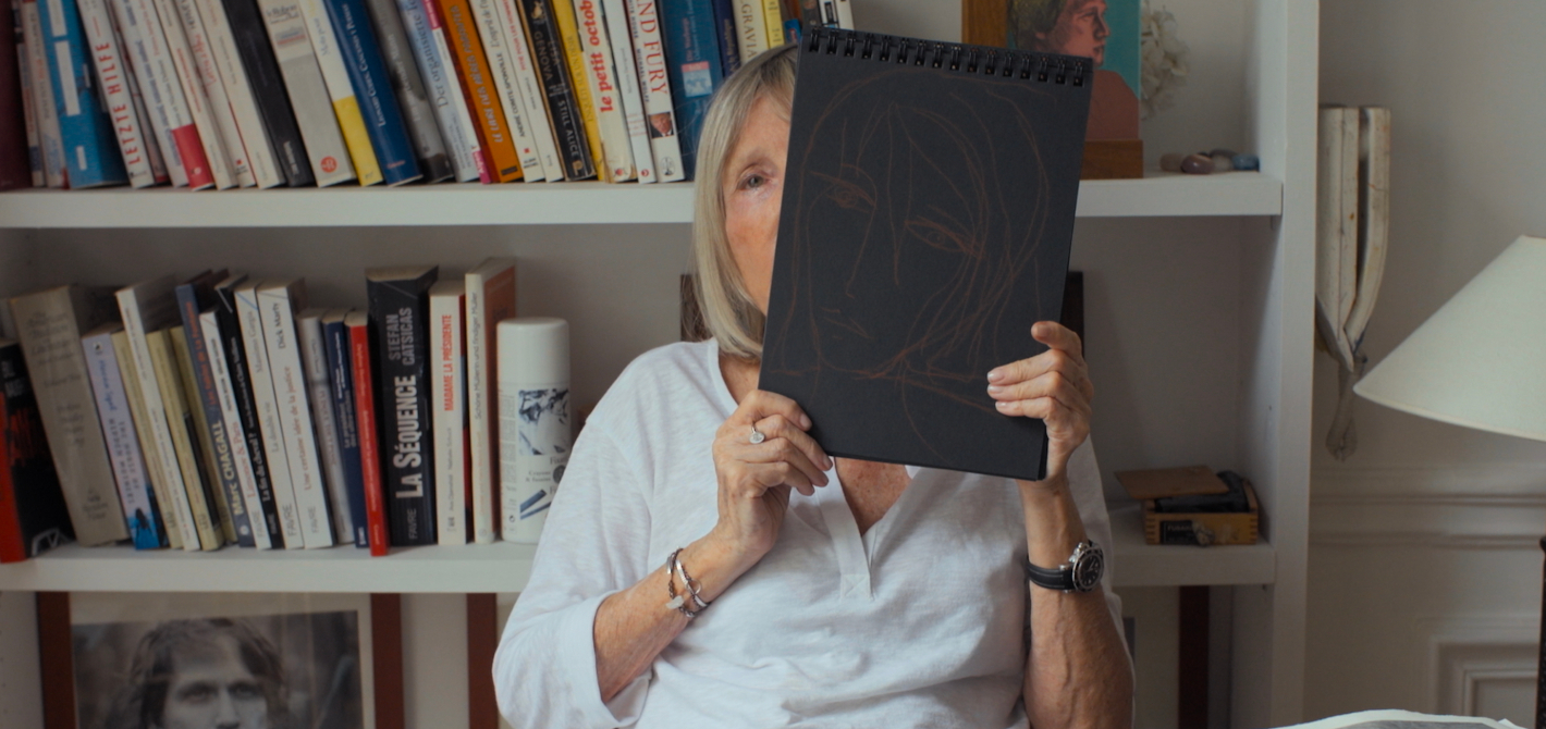 Starší žena s blond vlasy sedí ve své pracovně, drží před částí svého obličeje černé desky, na kterých je nakreslena tvář ženy. Za ní je knihovna s k knihami.