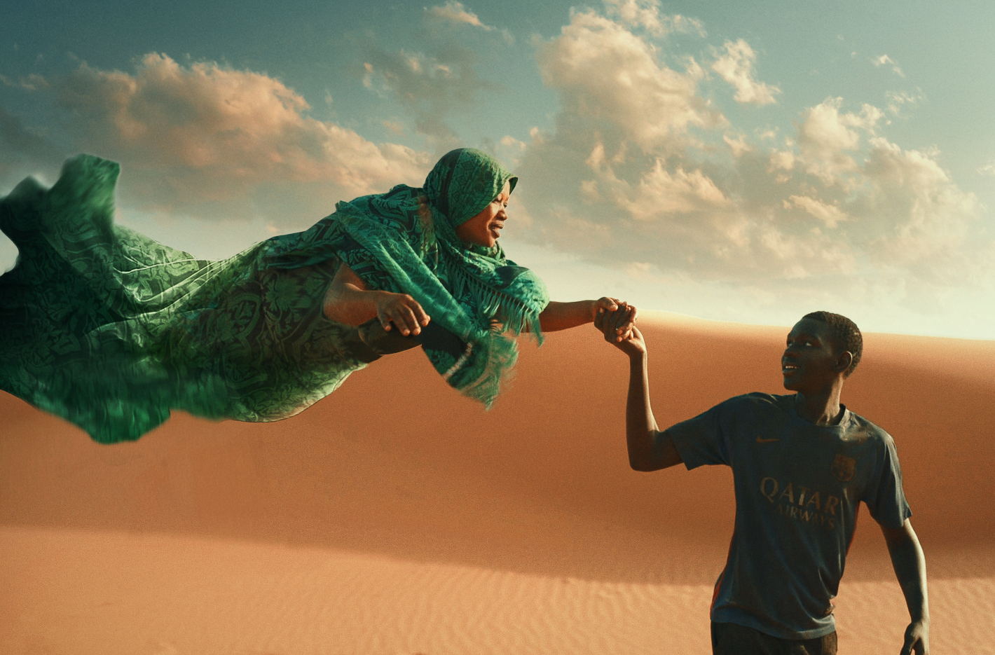 Mladý muž tmavé pleti drží za ruku ženu, která létá. Za nimi je písečná duna.