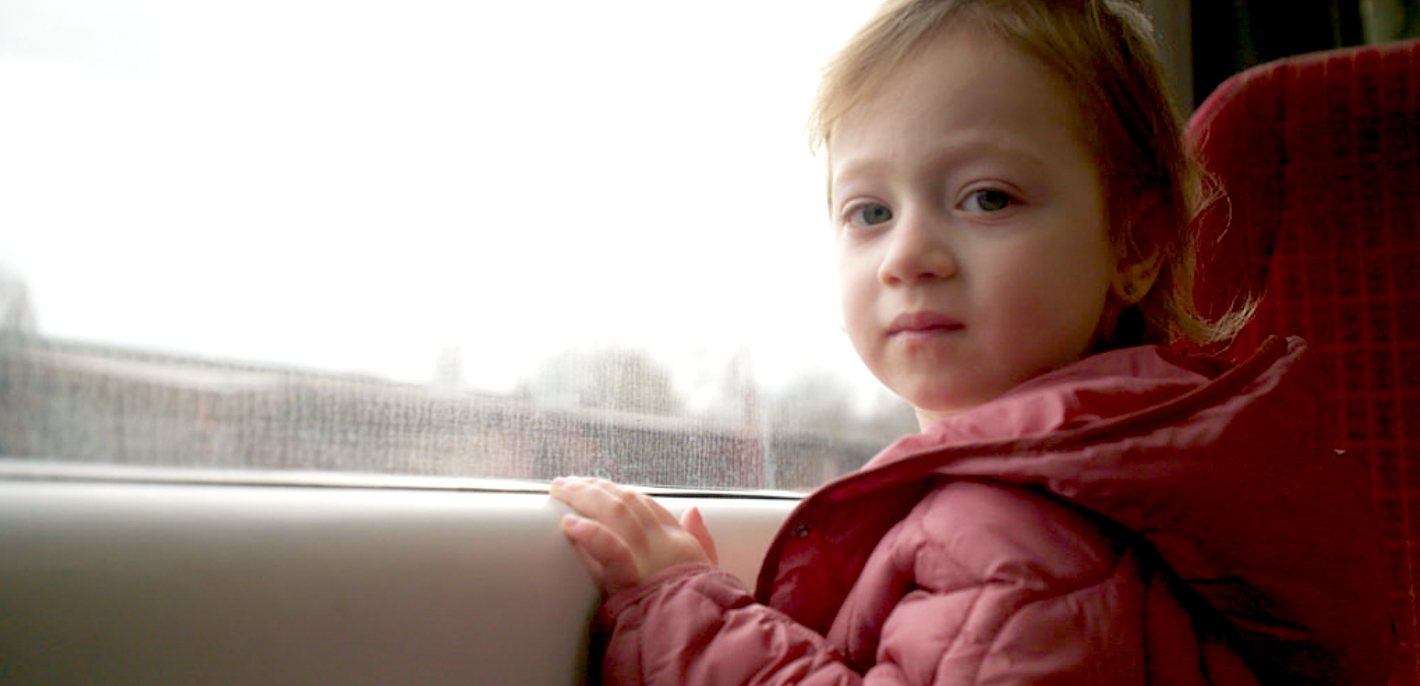 Malá holčička stojí u okna a drží se parapetu, dívá se do objektivu. 