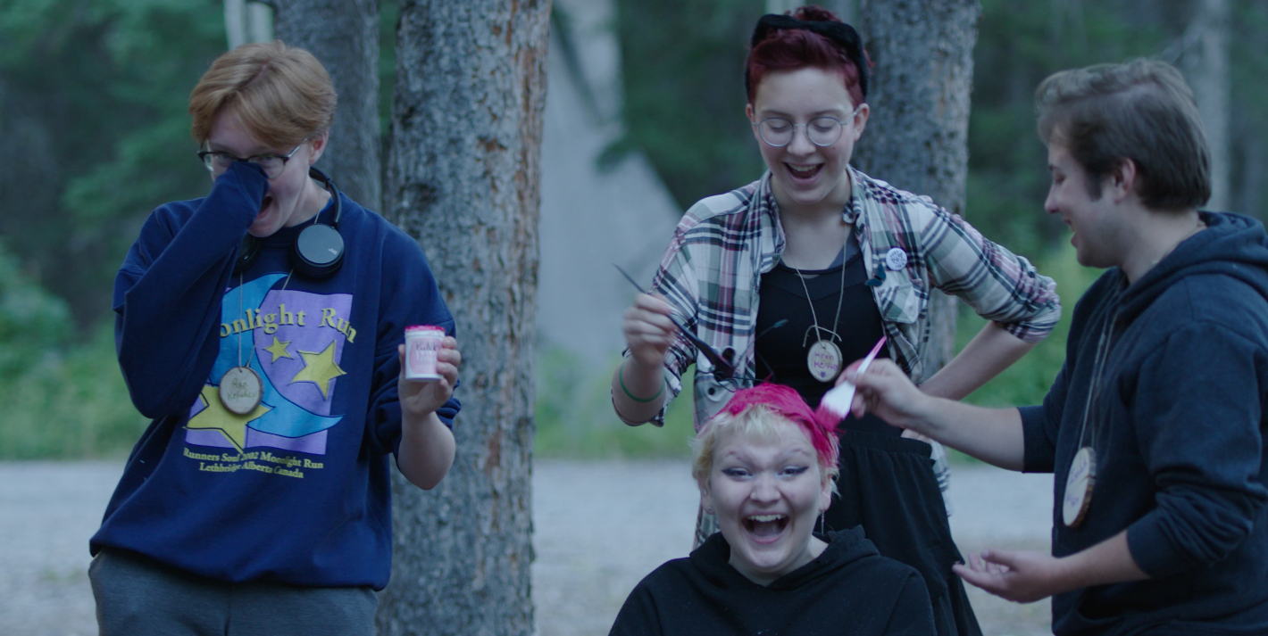 Čtyri mladí lidé se smějí, jedna dívka sedí a ostatní ji barbí vlasy na růžovo.