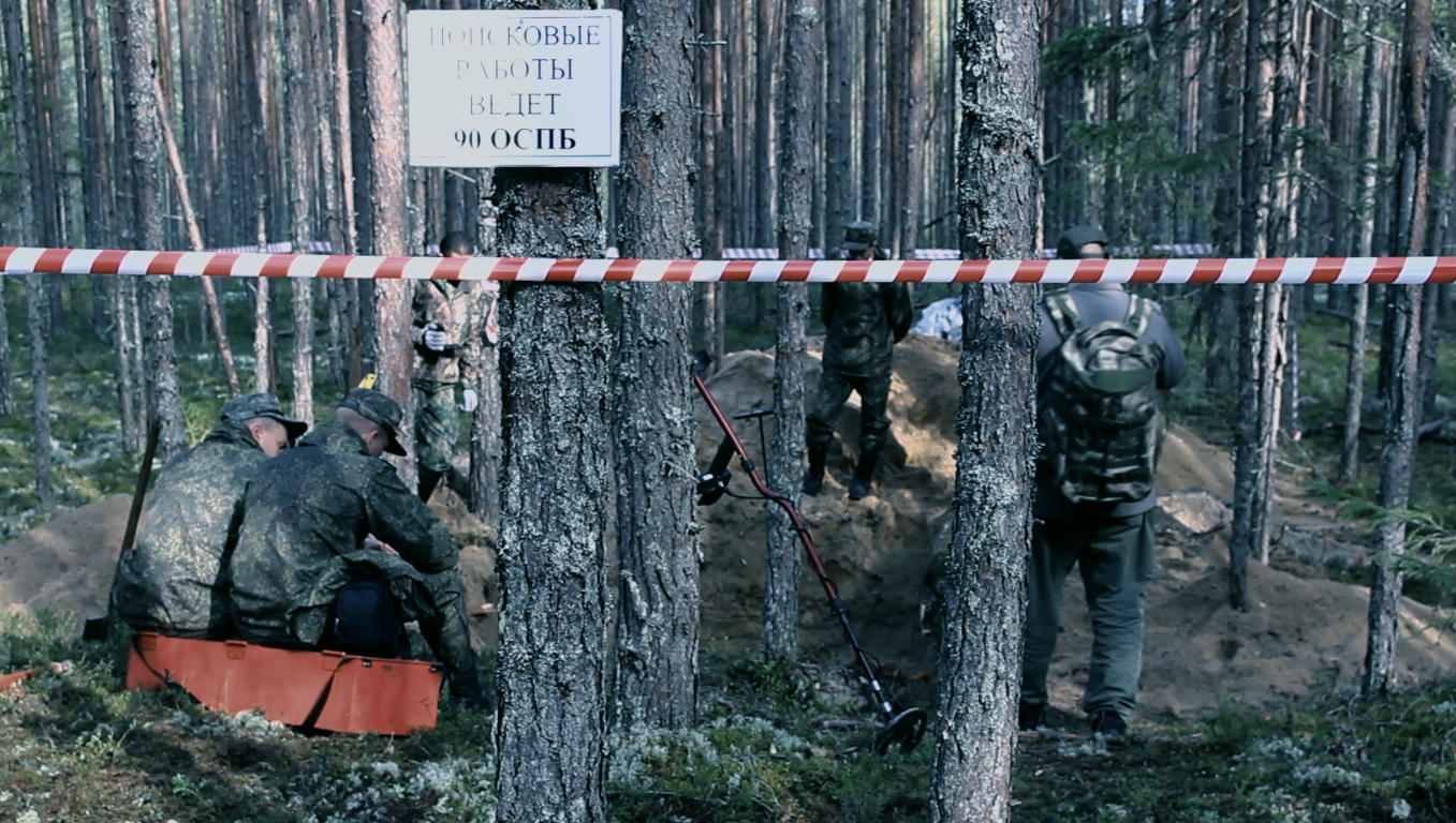 Muži v lese v maskáčích, před nimi vykopaná díra a u ní detektor kovů.