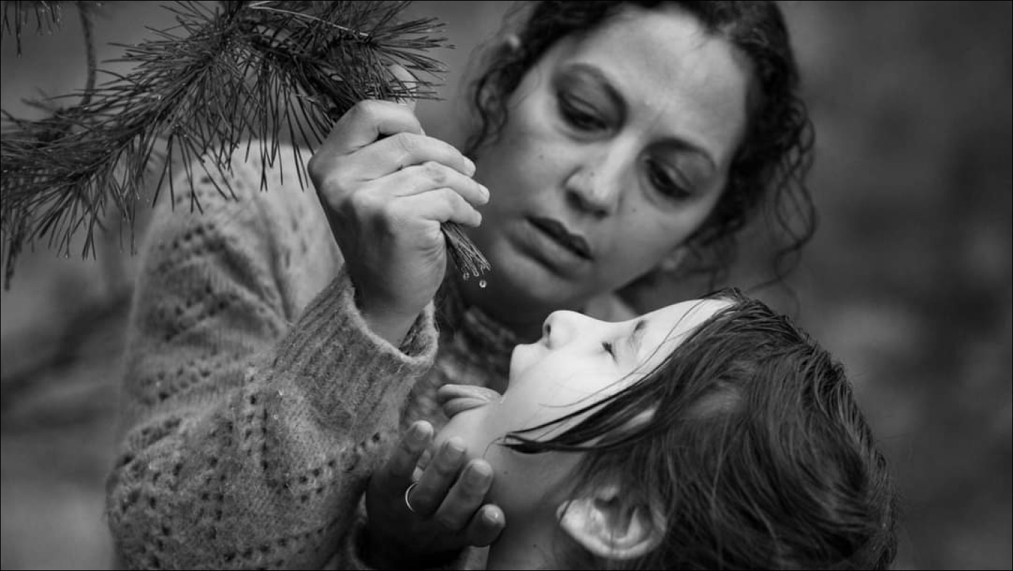 Černobílá fotka ženy, která drží větvičku borovice nad ústy dítěte, z větvičky stéká kapka vody.