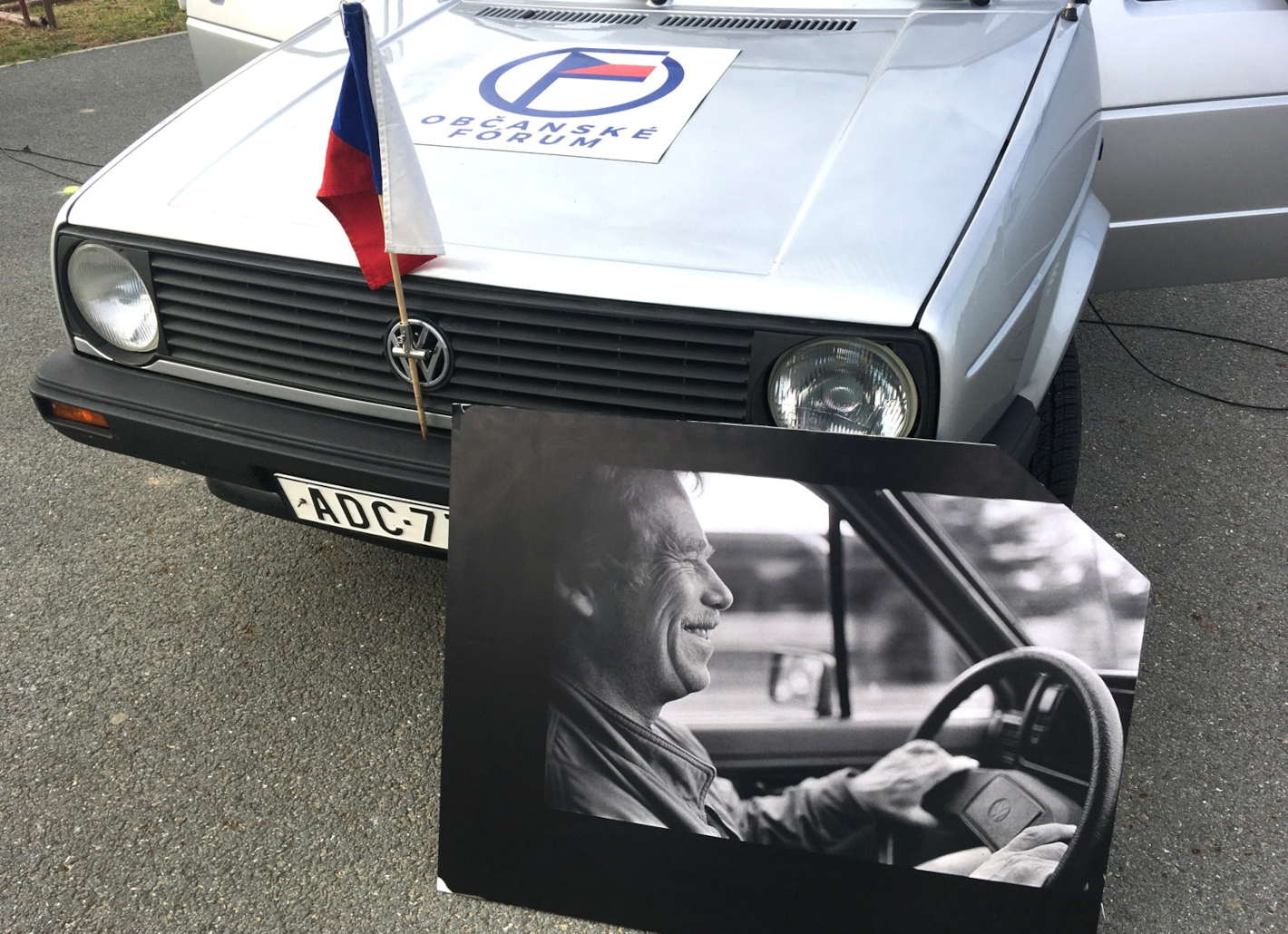 Auto VW Golf, na kapotě nálepka Občanské fórum, o přední sklo auta je opřená velká fotografie, na které je Václav Havel řídící toto auto.