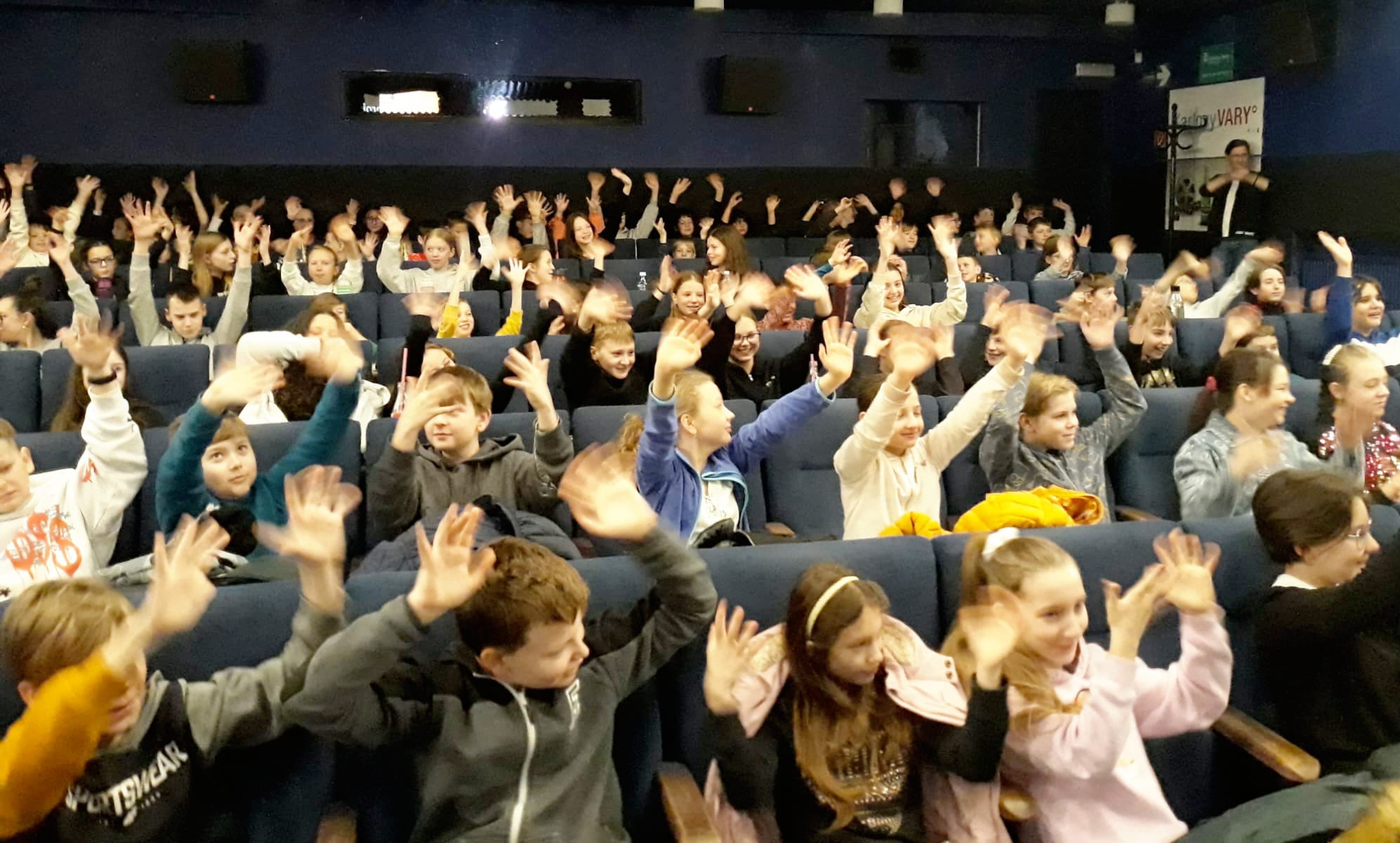 Plný sál kina, děti sedí, smějí se a mávají rukama.