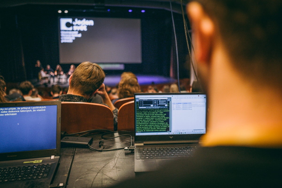 Pohled do sálu během panelové diskuze festivalu Jeden svět. V popředí jsou dva notebooky se spuštěným softwarem pro simultánní přepis mluveného slova do textu.