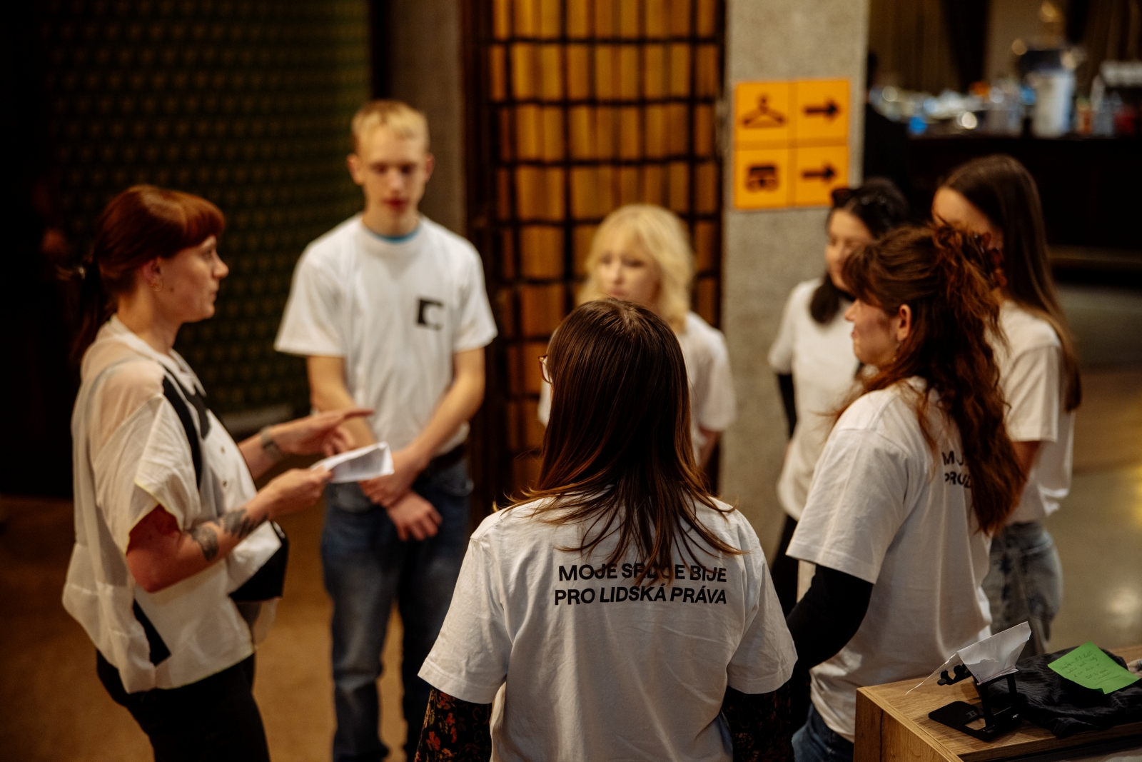 Sedm osob stojí v kruhu ve foyer, jedna z nich drží papír a vysvětluje pokyny.