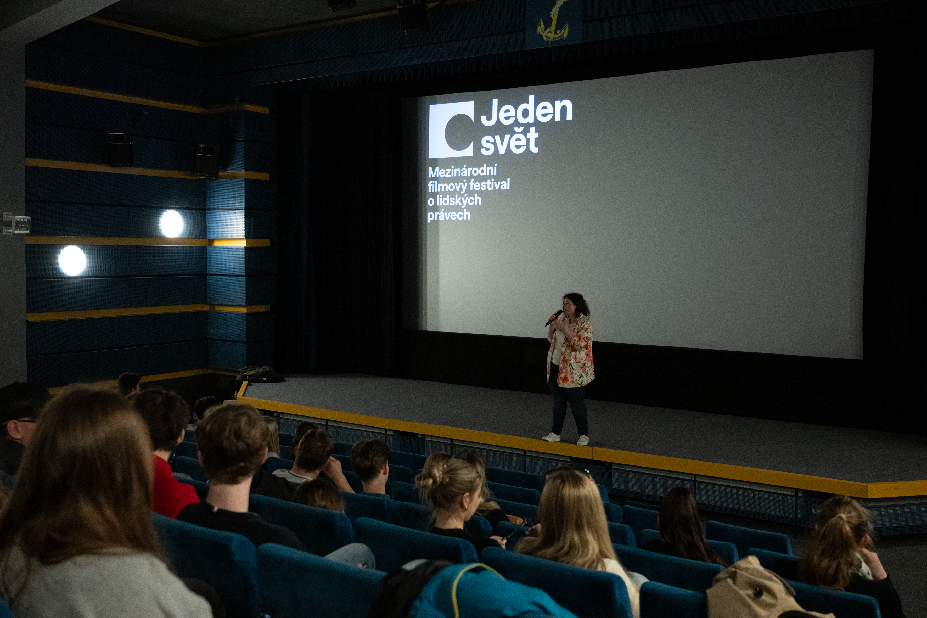 Moderátorka uvádí projekci pro školy v prostorech Biografu Kotva. V hledišti sedí žáci a žačky.
