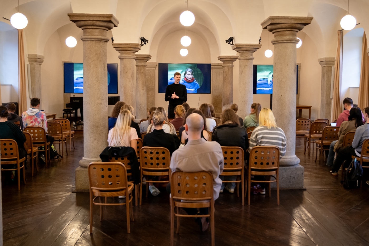 Přednáškový sál plný diváků. Vepředu stojí přednášející. Z ním filmové plátno.