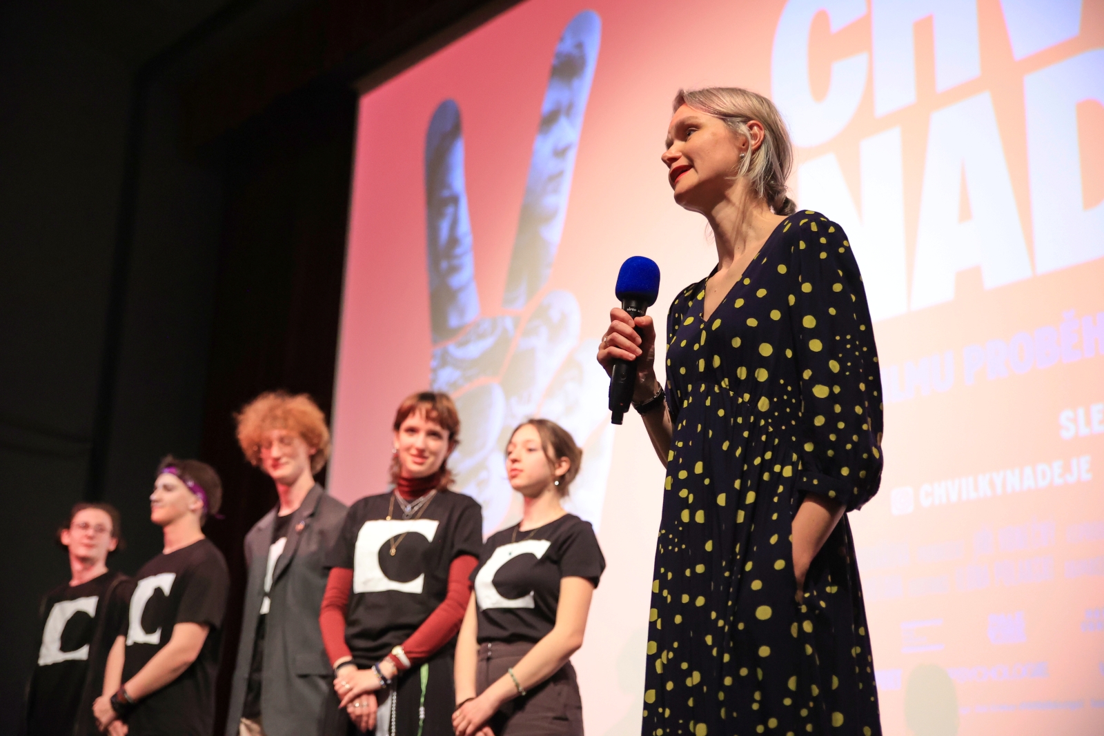 Skupina mladých lidí stojí na pódiu před filmovým plátnem, vedle nich žena s mikrofonem v ruce mluví k publiku.