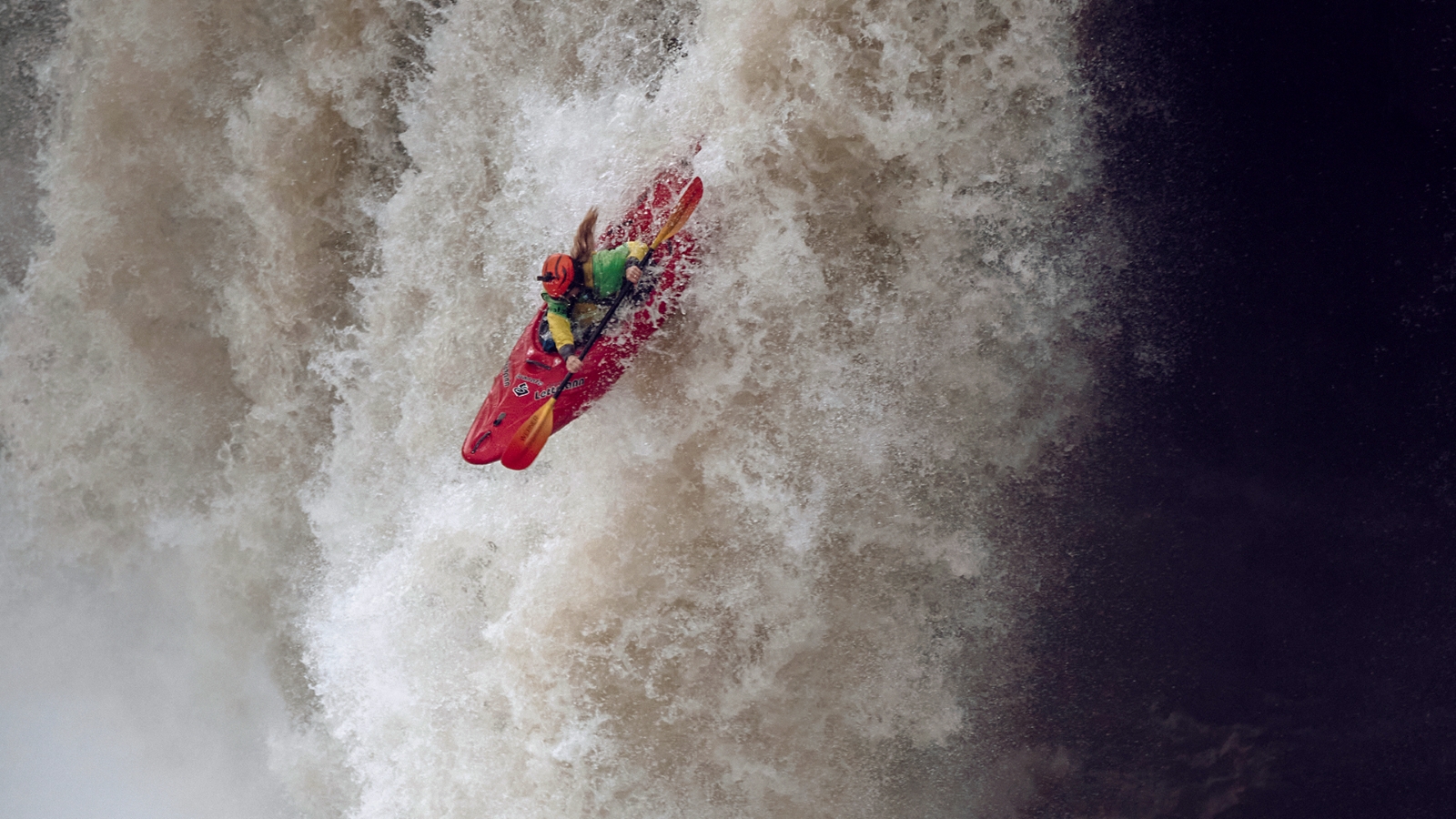 Kajakář sjíždí velký vodopád, obklopený stříkající vodou.