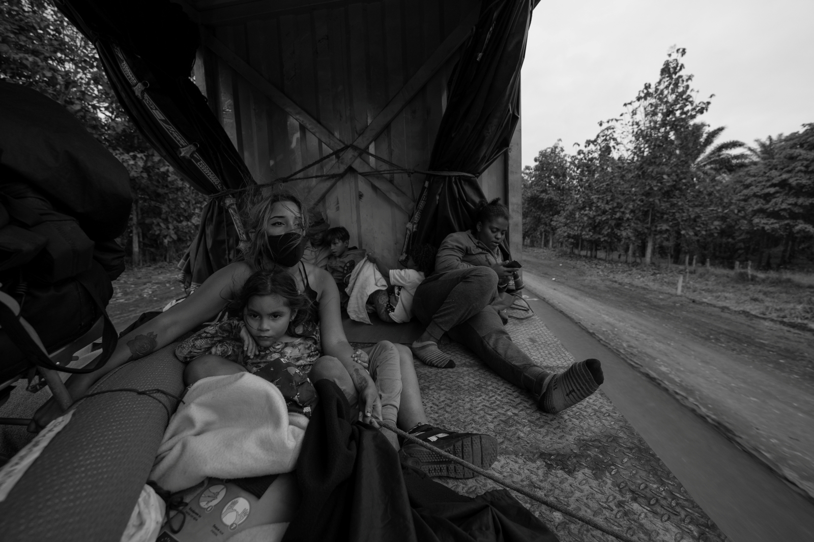Skupina lidí sedí na zadní části nákladního vozu, obklopená zavazadly, na venkovské cestě.