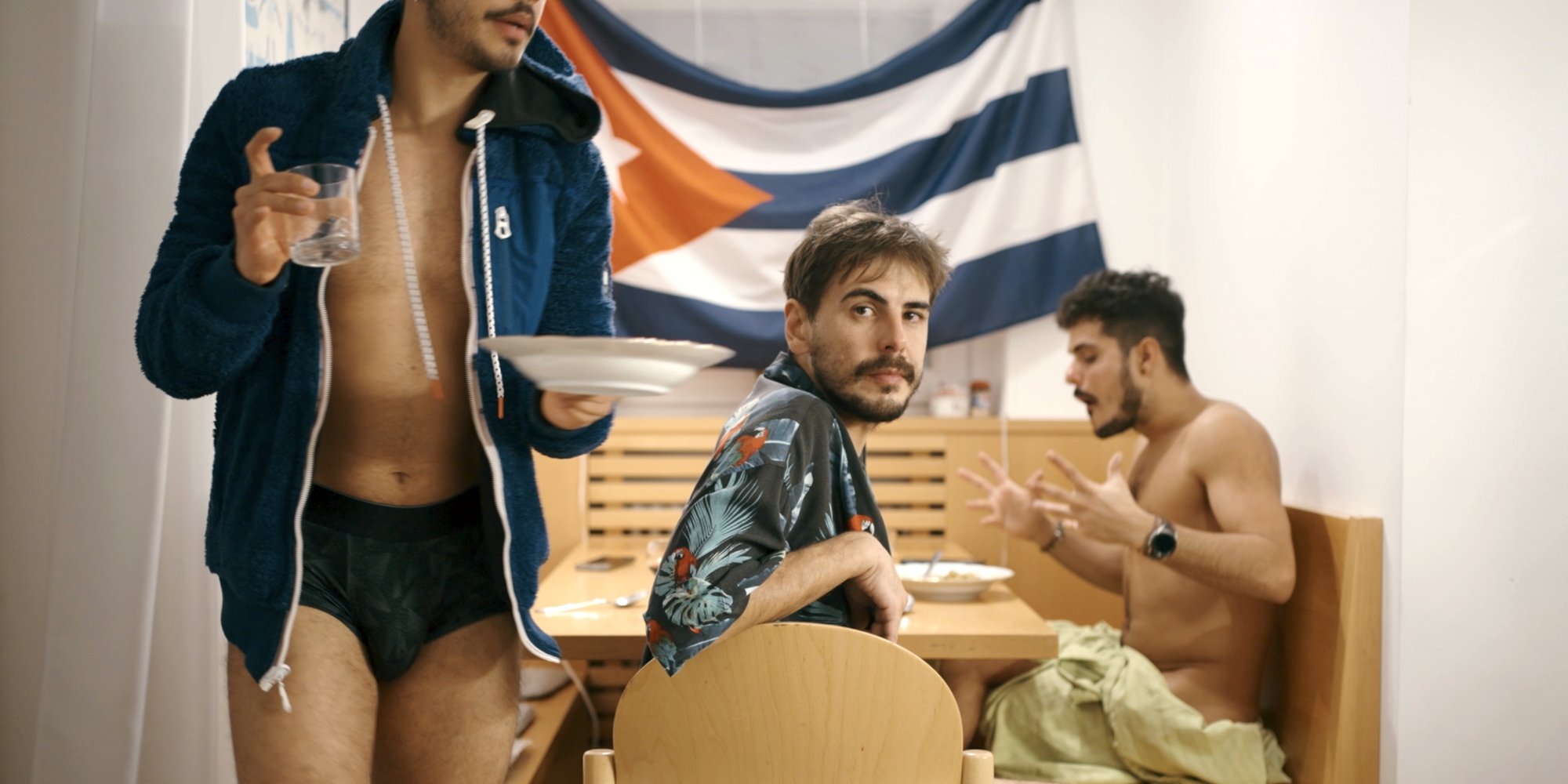 Dva muži sedí u stolu v interiéru u jídla, jeden odchází s talířem a sklenicí. V pozadí visí pruhovaná vlajka.