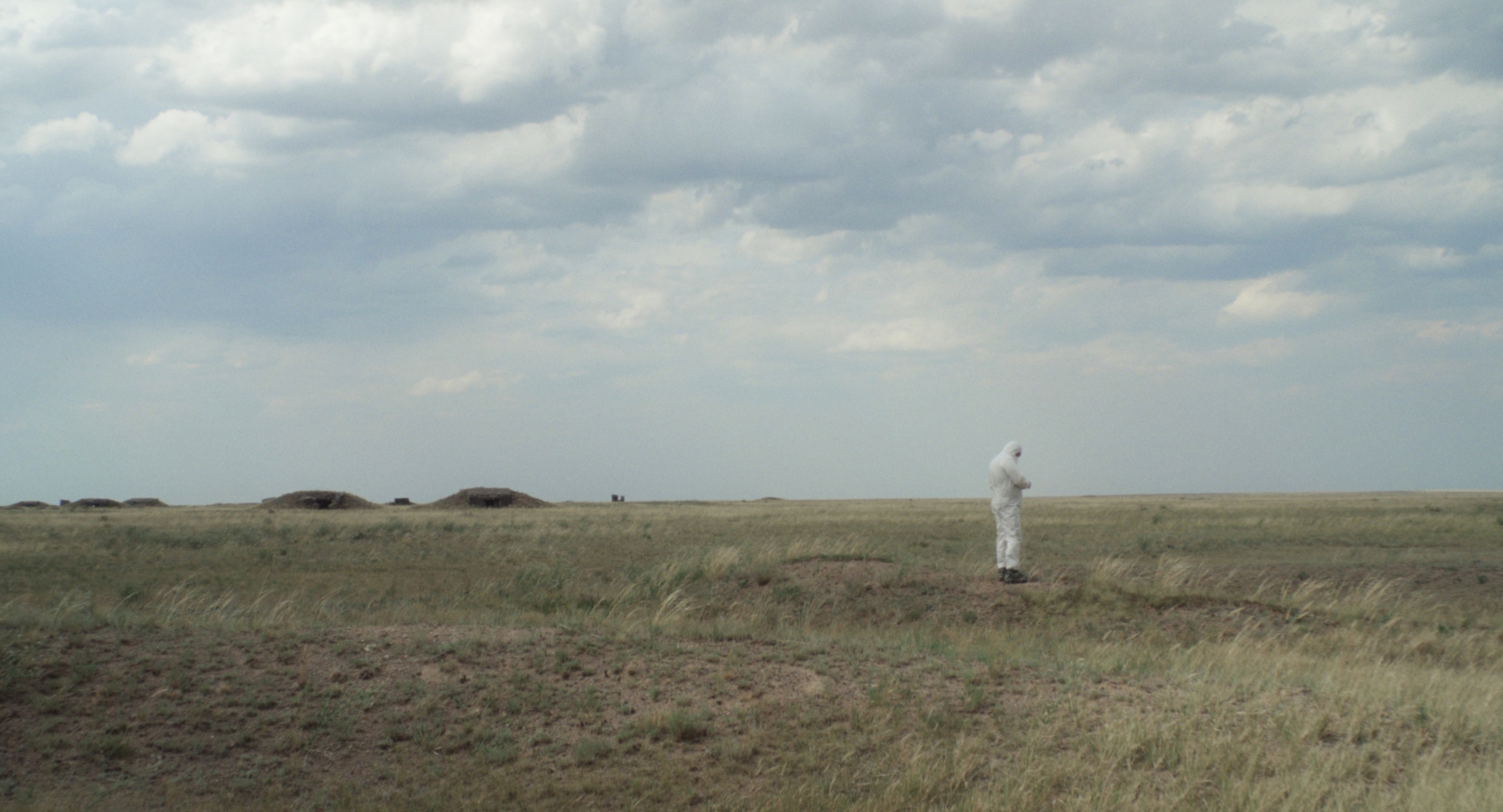 Osamělá postava v bílém oblečení stojí na rozlehlé travnaté pláni pod zamračenou oblohou.