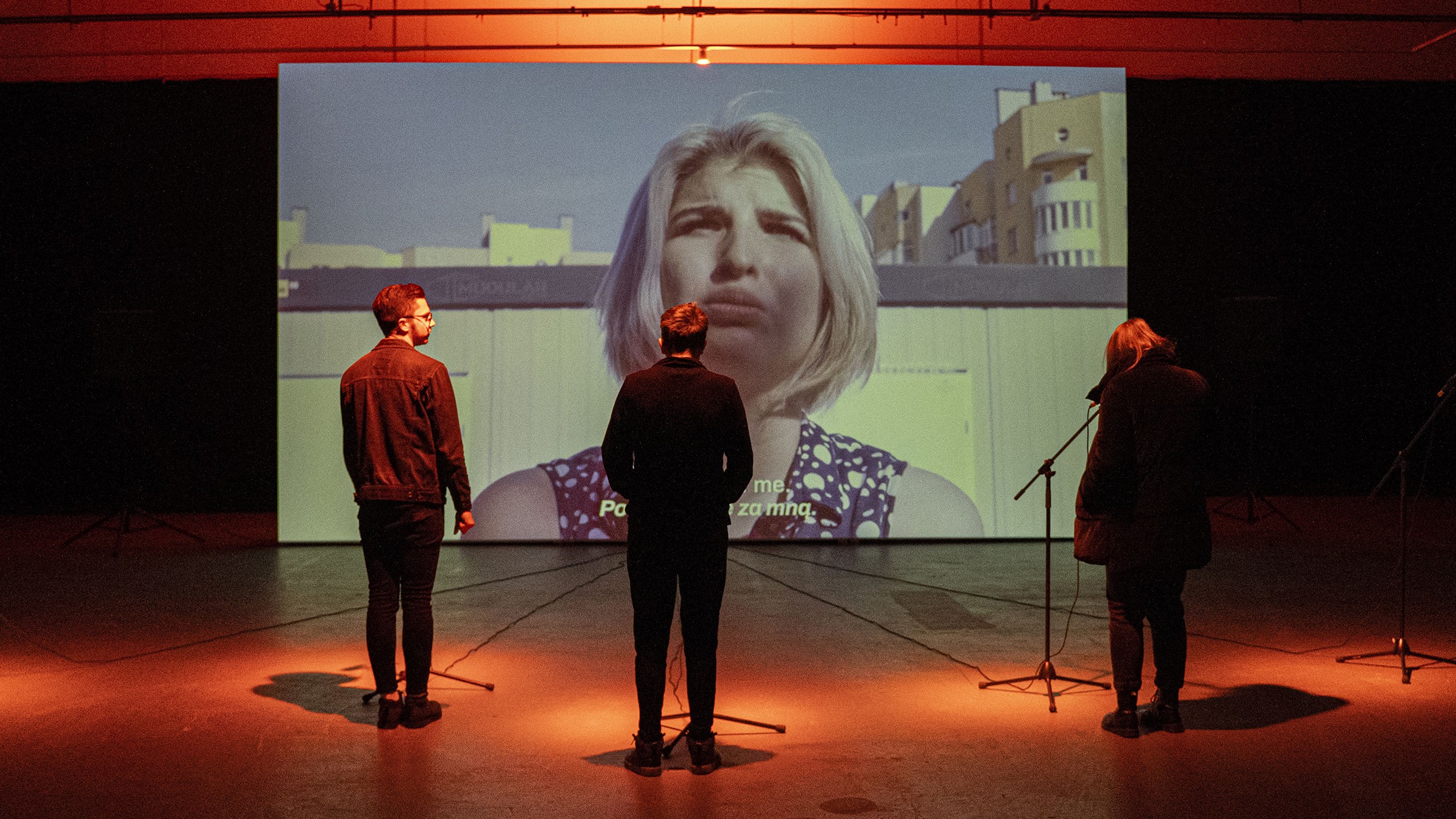 V tmavém sále stojí tři osoby před velkou projekcí ženy s blond vlasy; před každou osobou je mikrofon a prostor je nasvícen oranžovým světlem.