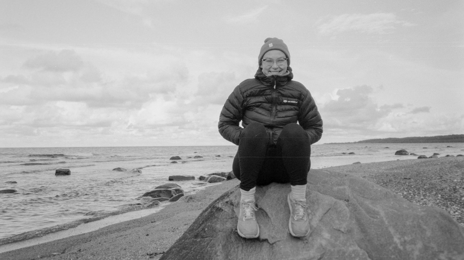 Žena v čepici a bundě sedí na kameni na pláži, za ní je vidět písek a moře.