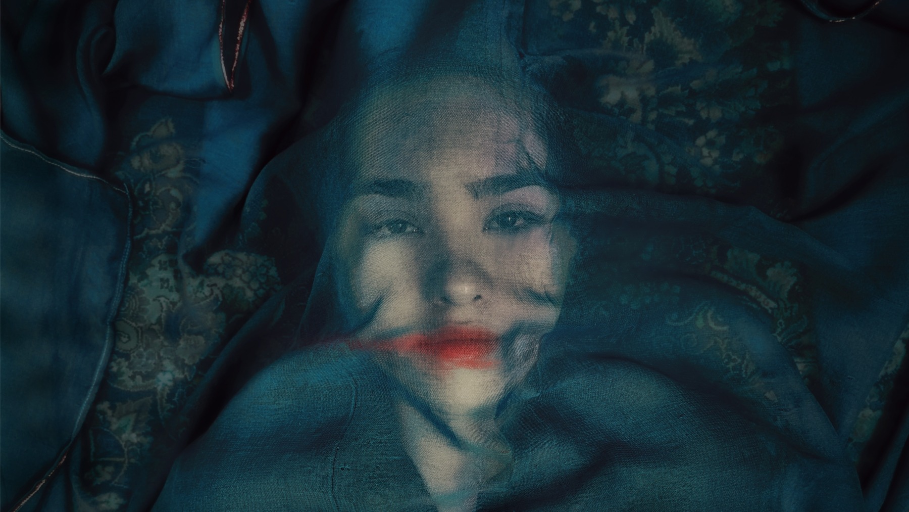 Ženský obličej viditelný přes modrou průsvitnou látku. Žena má výraznou červenou rtěnku rozmazanou kolem úst.