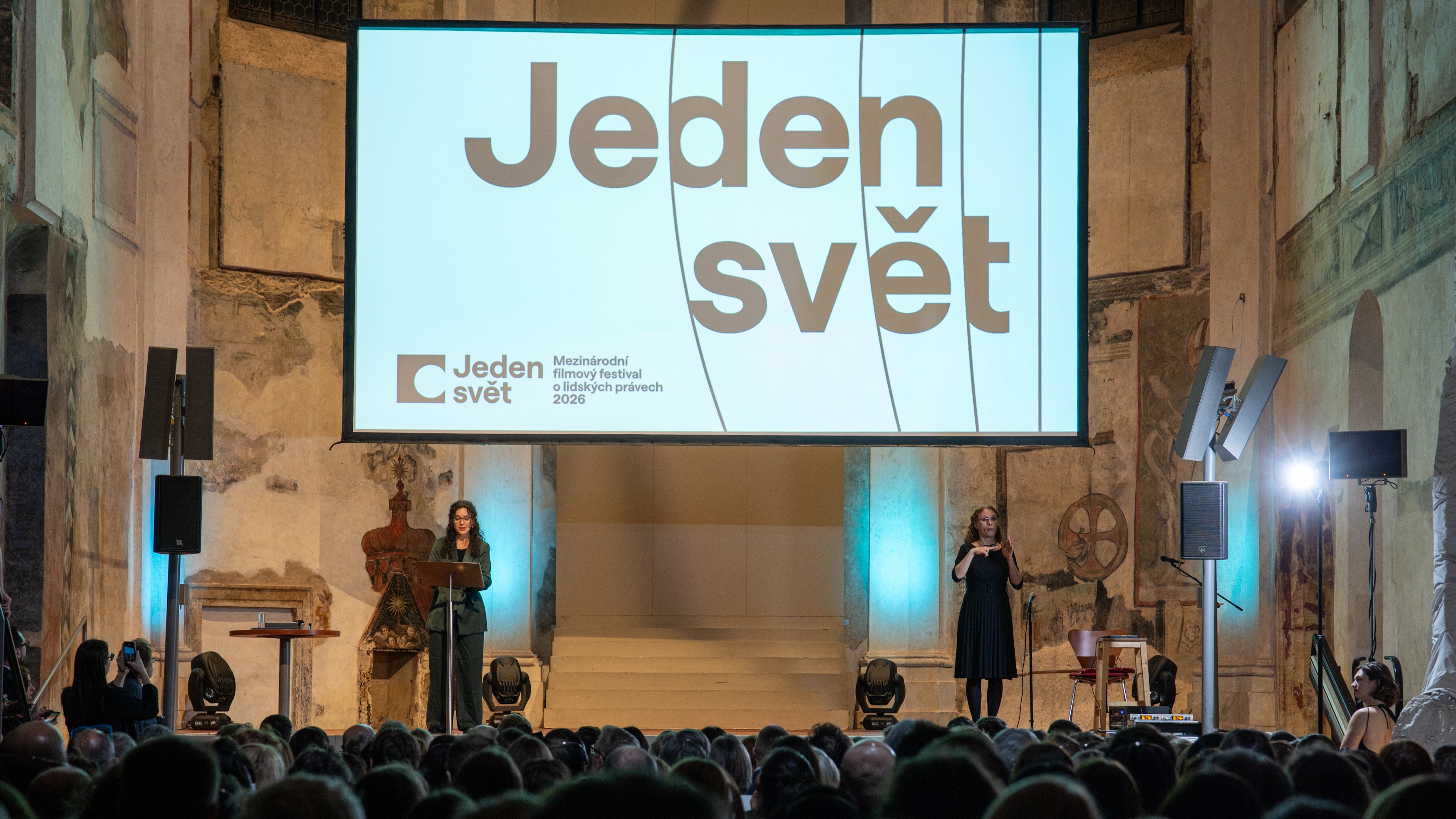 Na pódiu v historickém sále stojí řečnice u řečnického pultu a vedle ní tlumočnice do znakového jazyka. Za nimi je velké plátno s nápisem „Jeden svět – Mezinárodní filmový festival o lidských právech 2026“. Před pódiem sedí zaplněné publikum.
