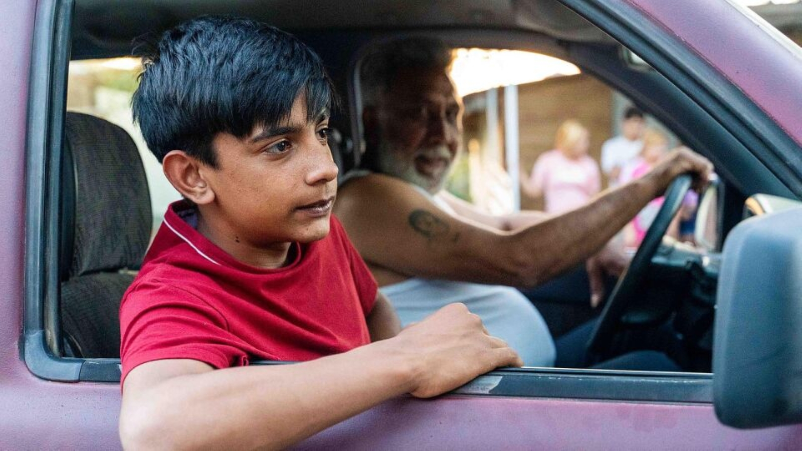 Chlapec sedí v autě a opírá se o otevřené okno auta a dívá se ven, zatímco starší muž sedí za volantem. V pozadí stojí několik rozmazaných postav před domem.