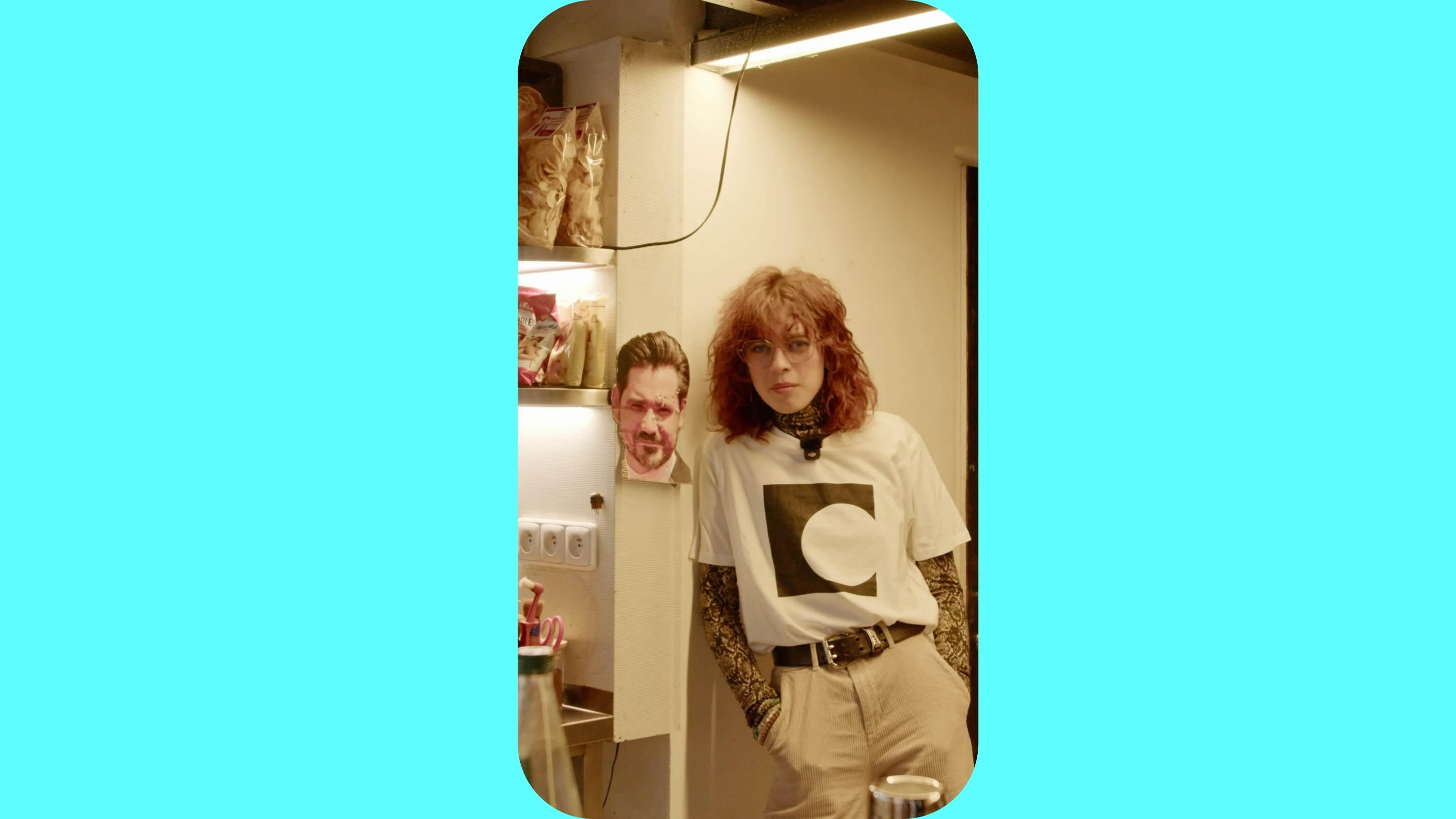 Žena se opírá ramenem o zeď, vedle ní visí na zdi mužská hlava vytištěná na papíře.