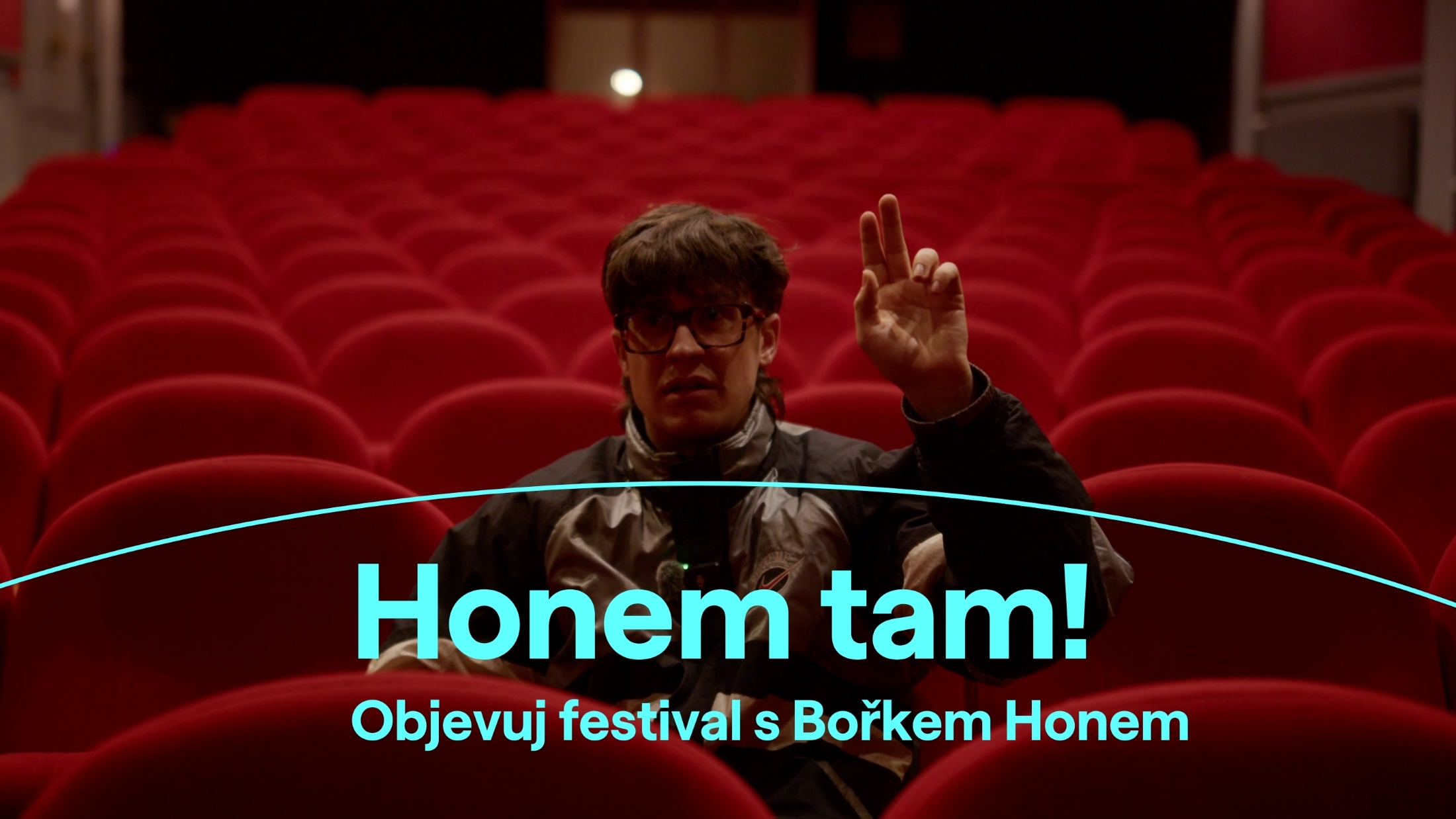 Muž sedí v hledišti a zvedá ruku jako kdyby se hlásil.