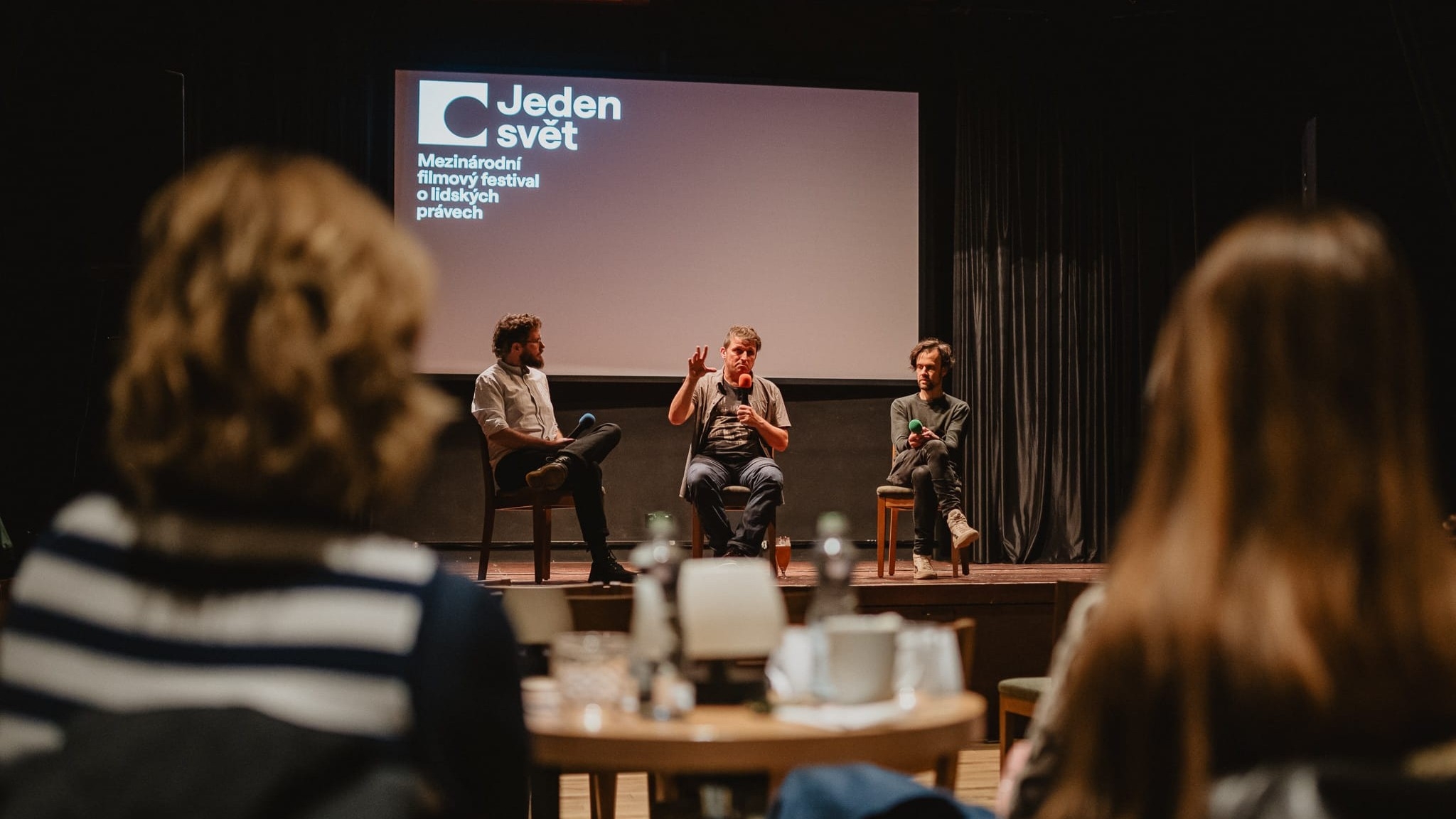 V sále před filmovým plátnem debatují tři muži. Za nimi je logo festivalu Jeden svět.