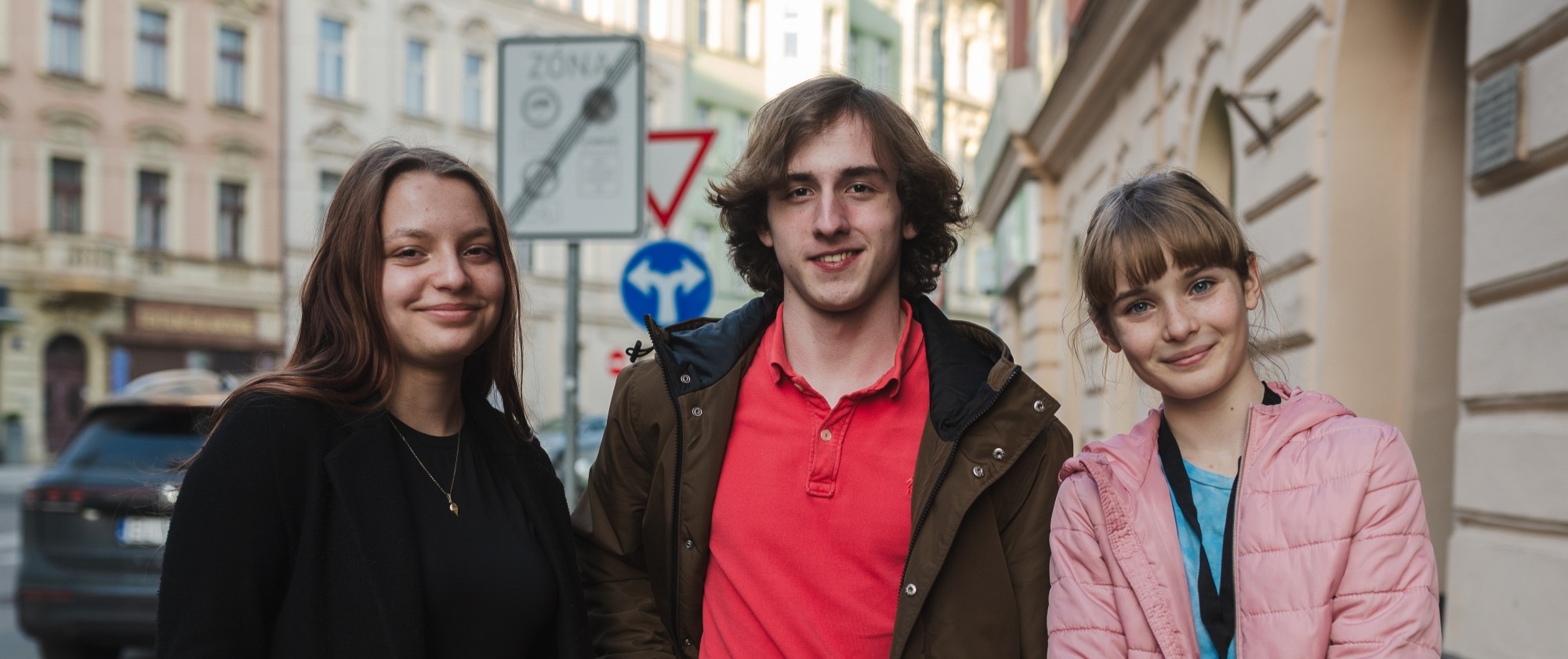 Tři osoby stojí na ulici vedle sebe. Za nimi jsou vidět činžovní domy a dopravní značky.