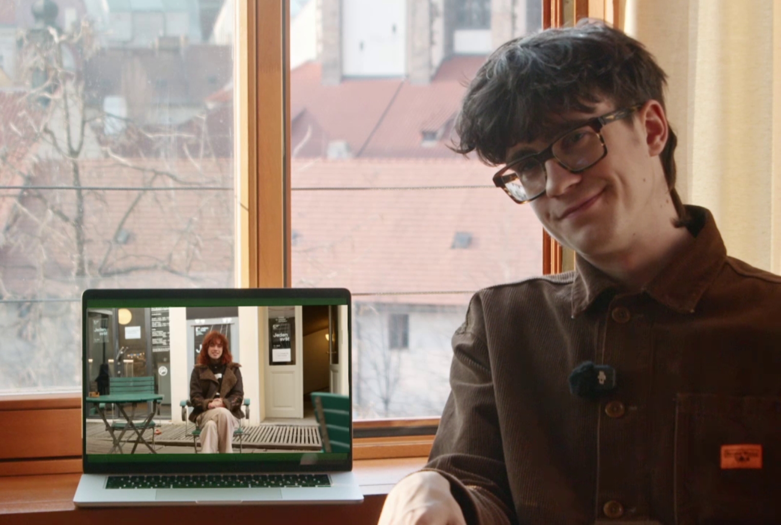 Muž sedí u okna s výhledem na kostel a vedle sebe má položený notebook, na které je video ženy sedící na židli na dvorku.