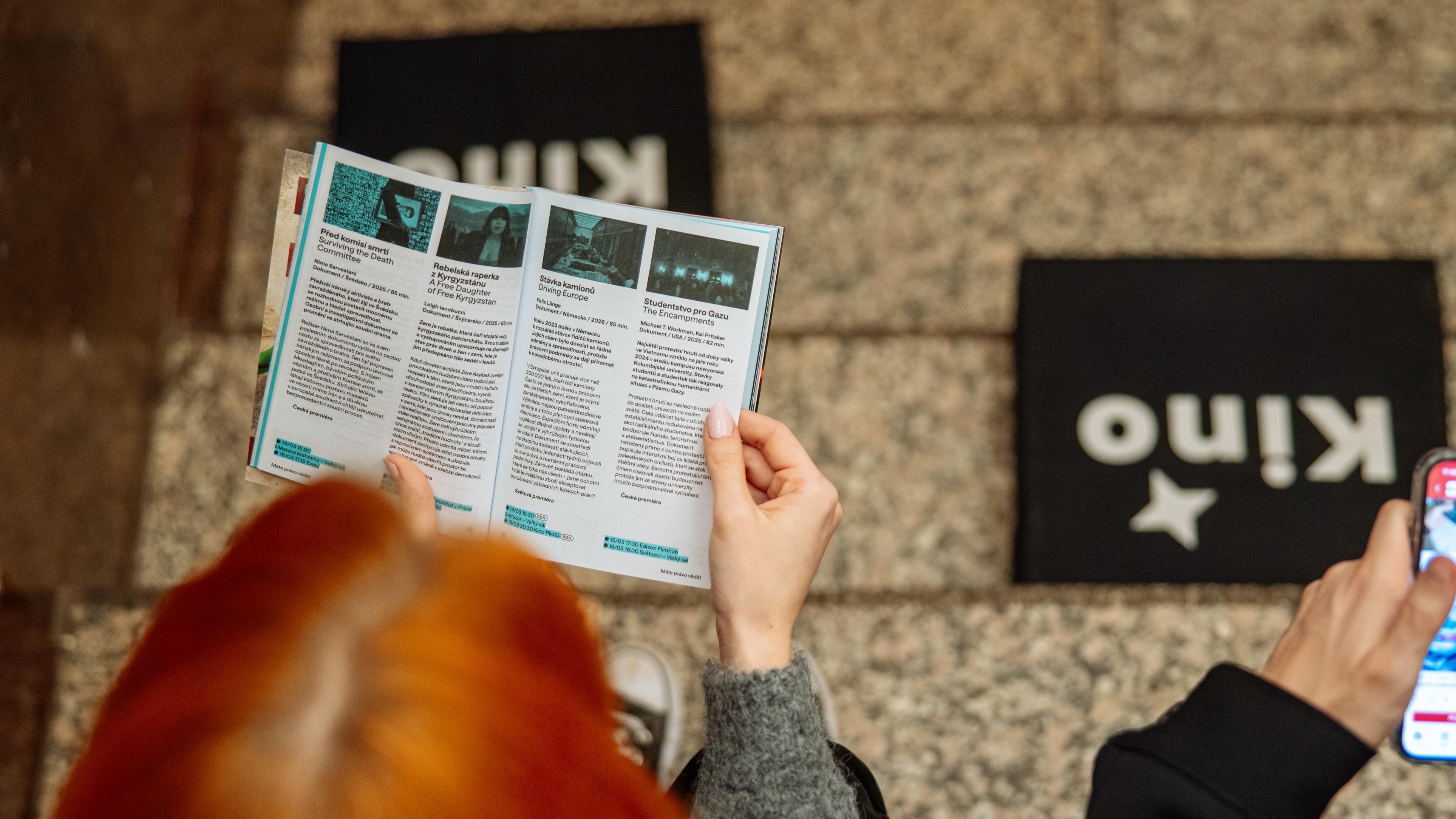 Žena sedí na schodech a drží otevřený tištěný program s přehledem fotek filmů a textů, na schodech leží polštáře s nápisem Kino.