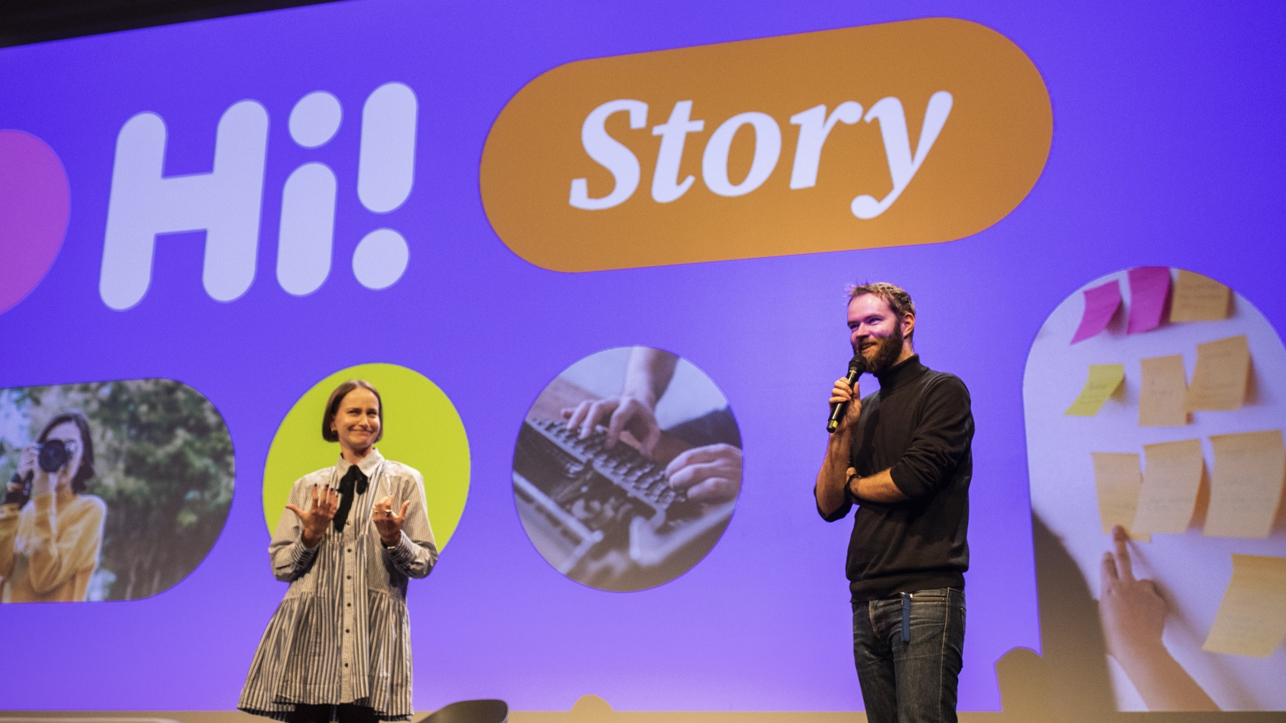 Na pódiu stojí dva přednášející před velkou projekční plochou s nápisem „Hi! Story“ a několika fotografiemi. 