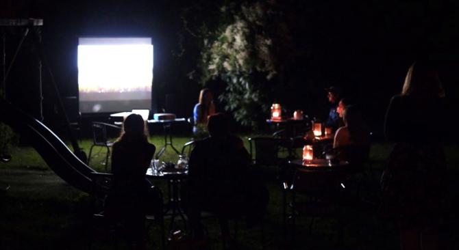 Lidi sedí večer na zahradě u stolečků a sledují promítání filmu