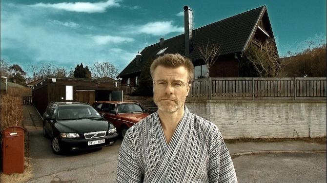Uprostřed klidné obytné ulice stojí muž středního věku s krátkými vlasy, brýlemi a krátkým vousem, oblečený ve světlém županu s tmavým vzorem. Za ním je rodinný dům, dvě zaparkovaná auta v příjezdové cestě a červená poštovní schránka u chodníku.