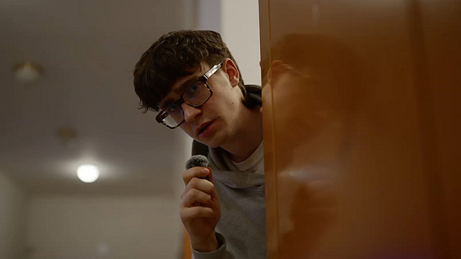 Mladý muž s brýlemi vykukuje zpoza dveří a drží v ruce malý mikrofon.